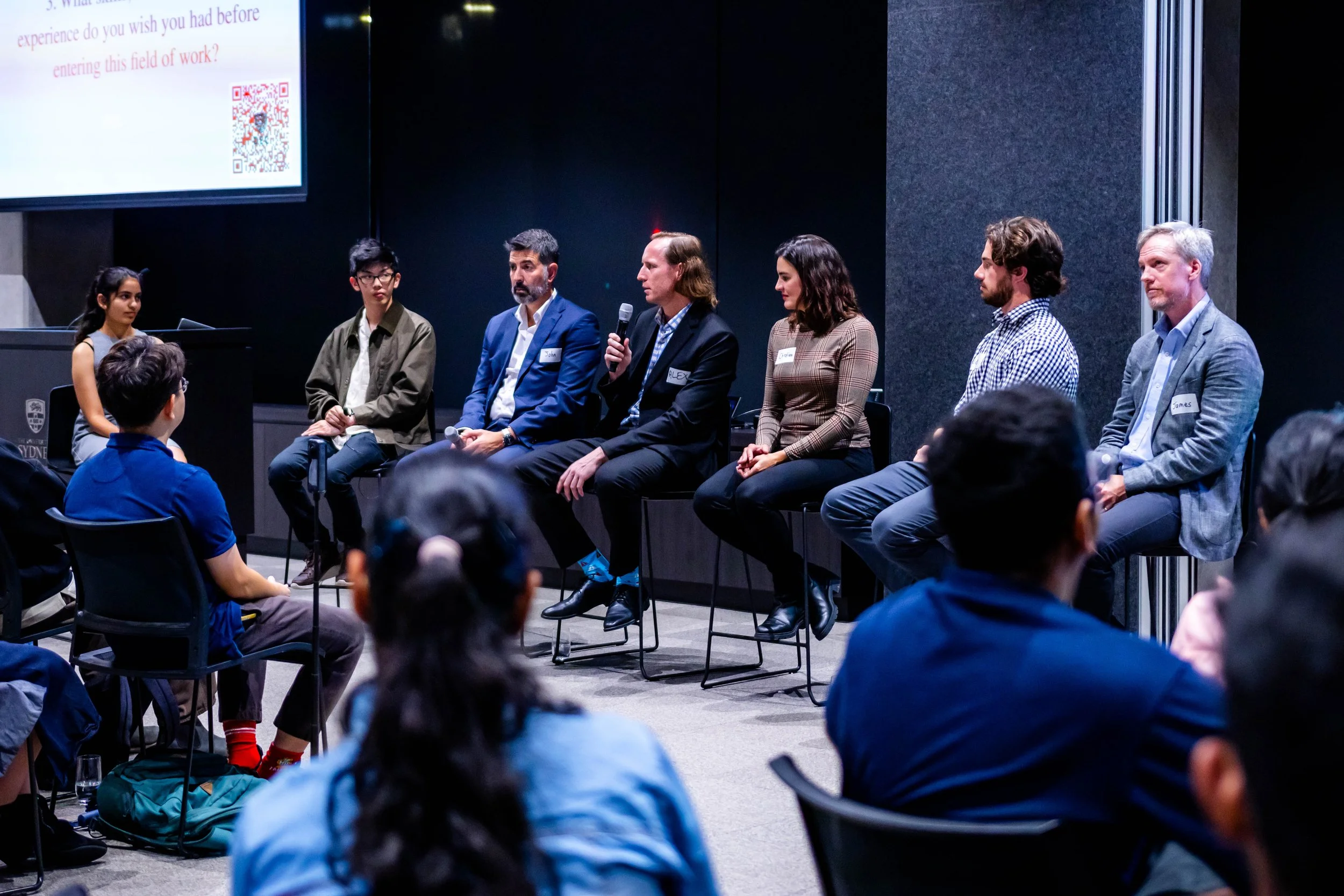 Panel discussion with seven people seated on stage, one woman holding a microphone, audience members seated facing the panel, and a large screen displaying text and a QR code in the background.