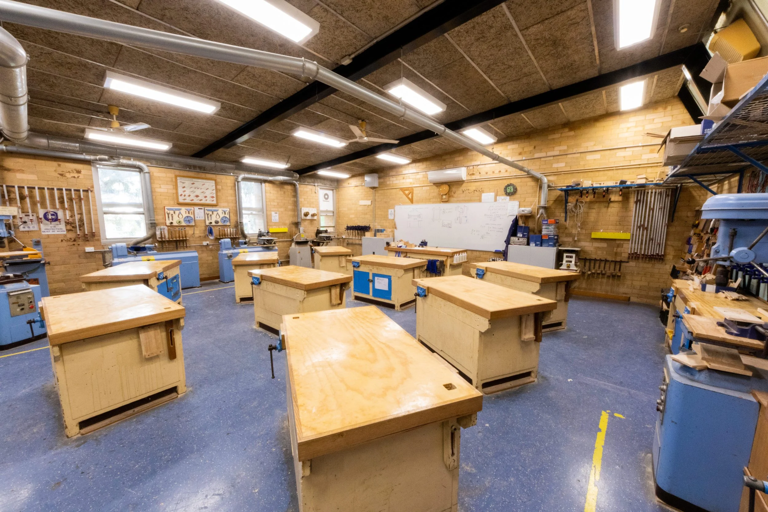 Woodworking classroom with multiple workbenches, tools, and equipment along the walls, whiteboard at the front, and fluorescent lighting.