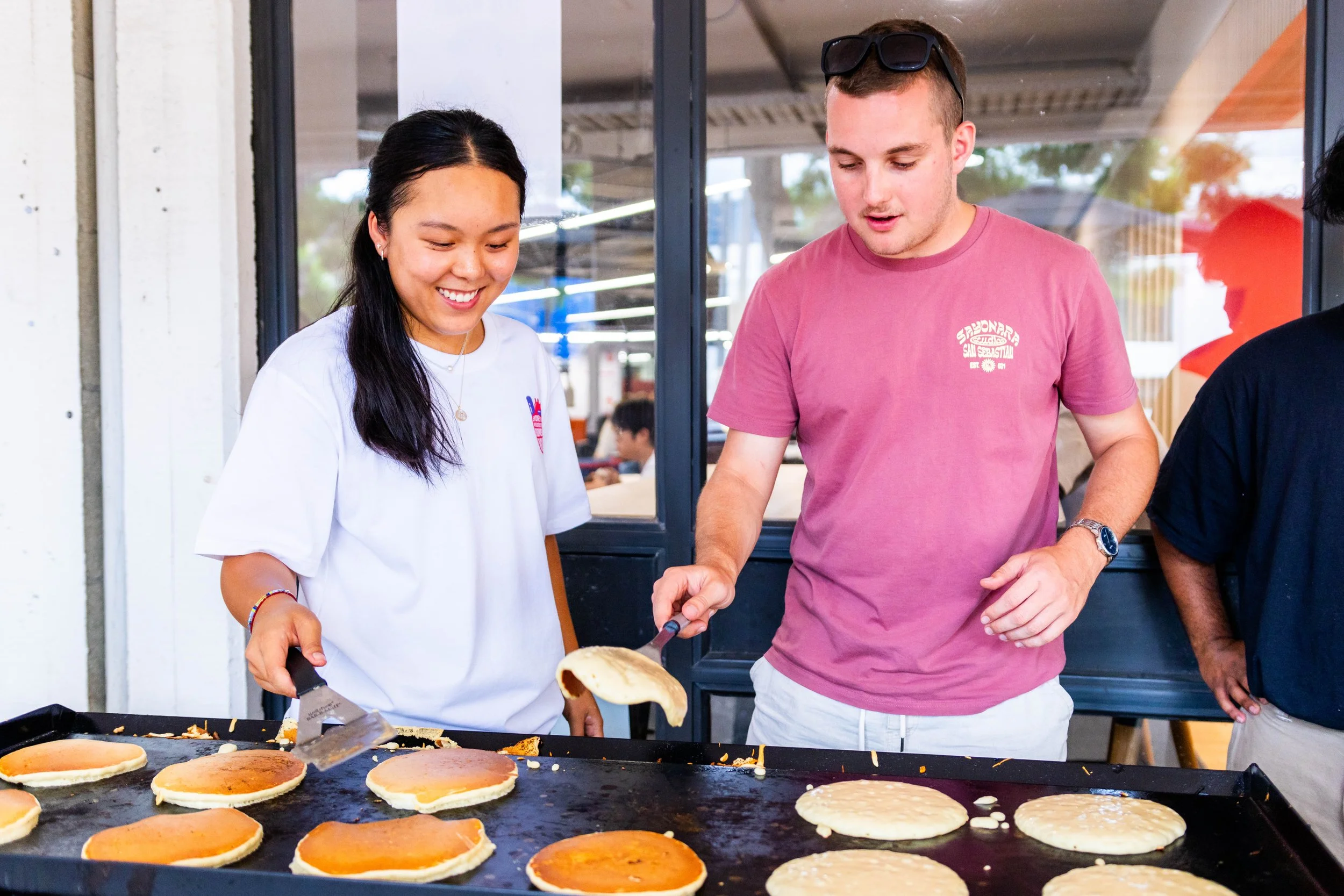 A woman and a man cooking pancakes on a flat griddle at a restaurant or cafe, with the woman smiling and the man flipping a pancake.