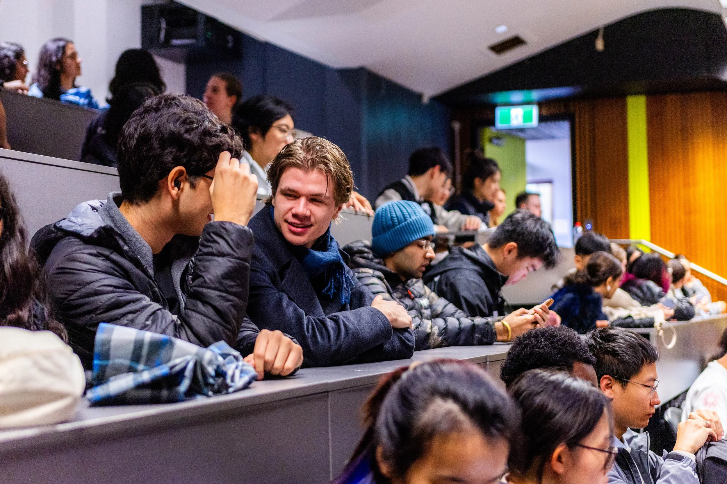 Students sitting in an auditorium, engaging in conversation and using their phones, during a lecture or event.
