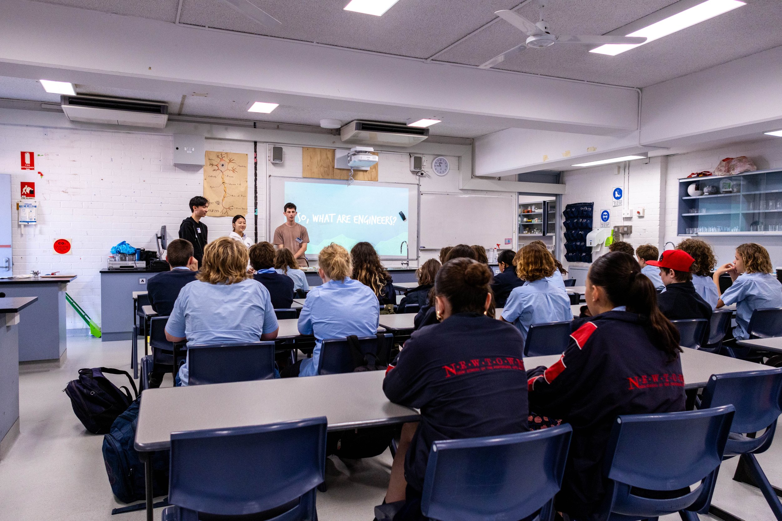 Students in a classroom attending a presentation about engineering, with a projector displaying 'So, what are engineers?' on the whiteboard.