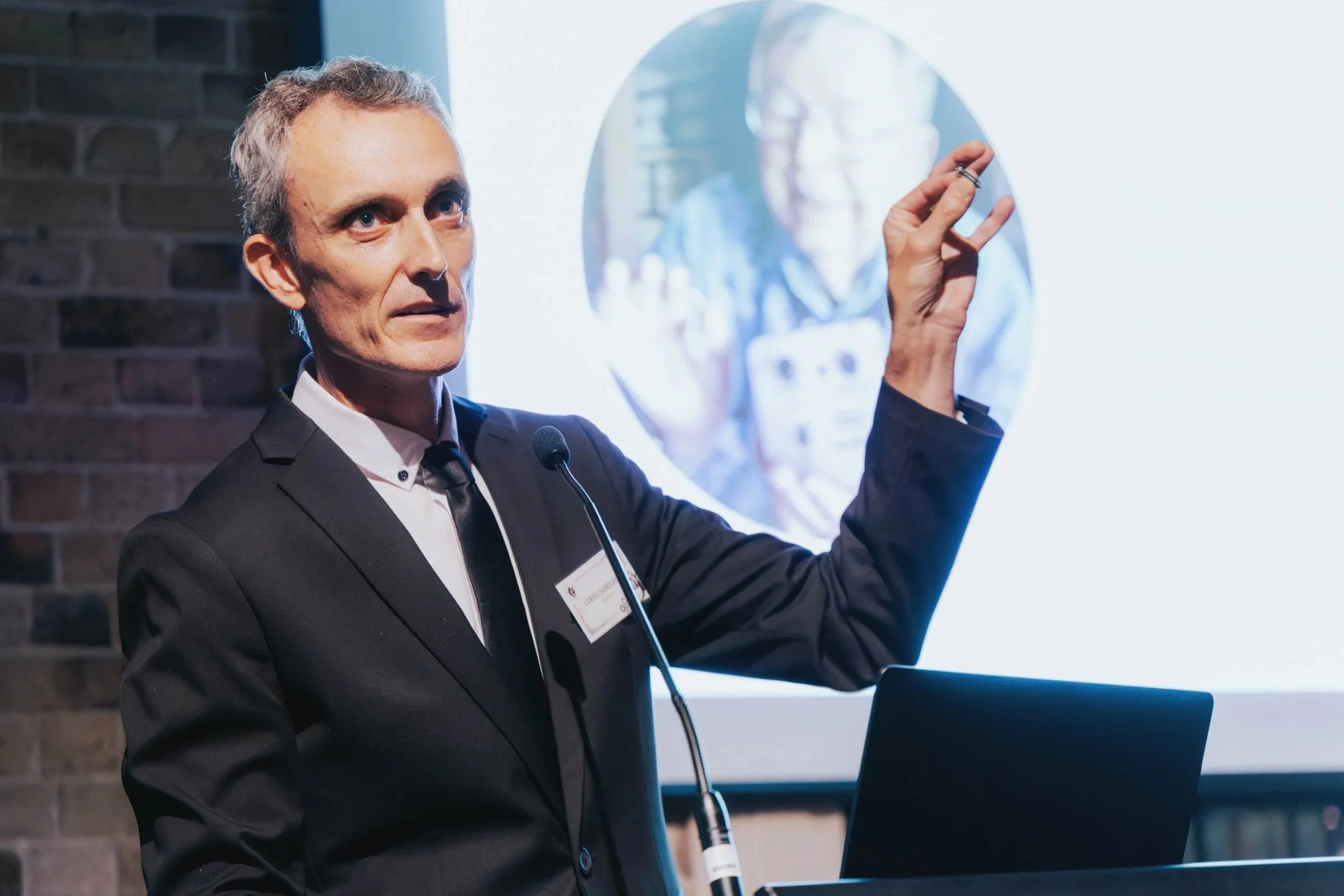 A man in a suit giving a presentation in front of a screen, gesturing with his hand, with a woman’s face visible on the screen in the background.