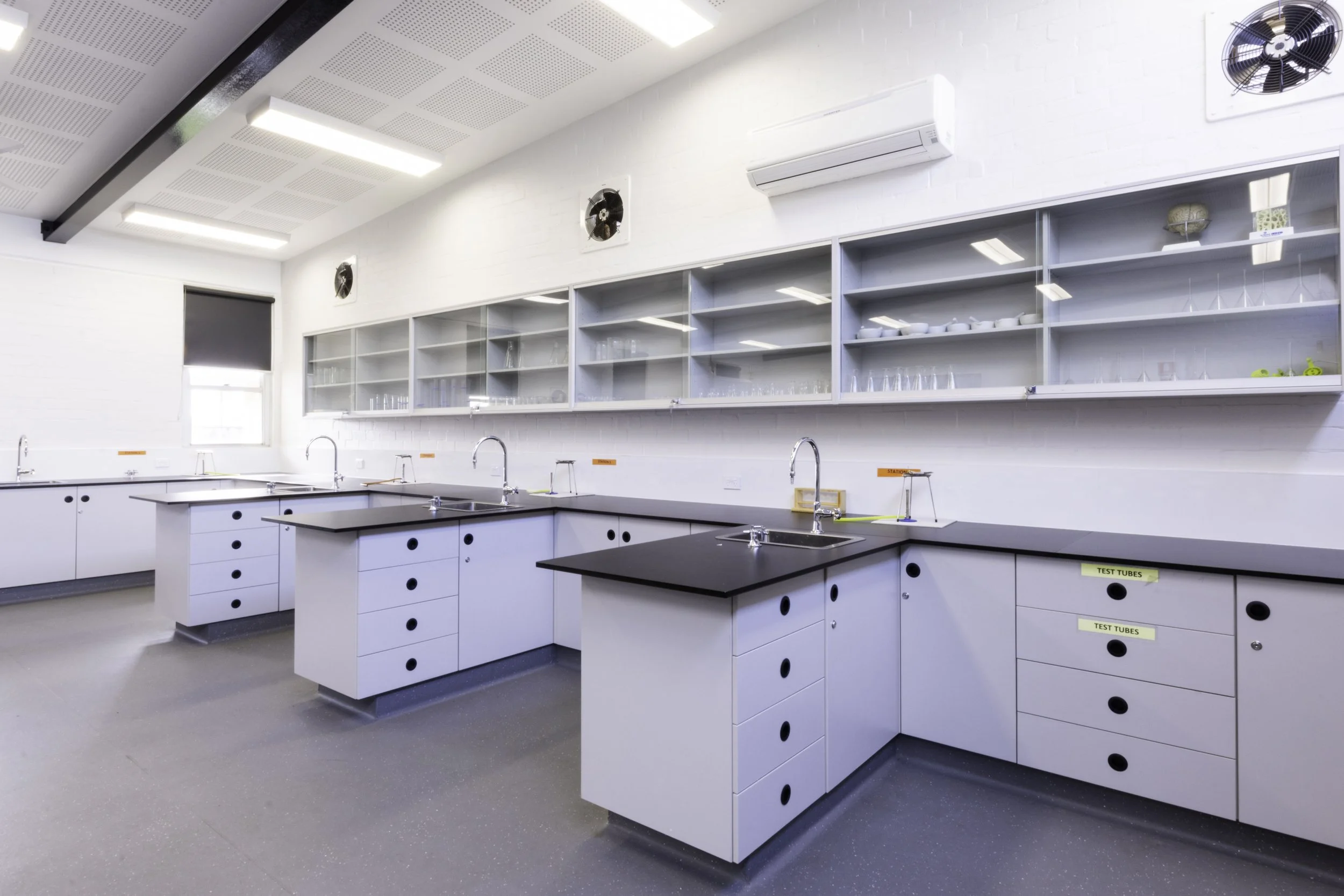 A clean, modern laboratory with white cabinets, black countertops, multiple sinks, and open shelves with laboratory equipment. There is a window and an air conditioning unit on the wall.