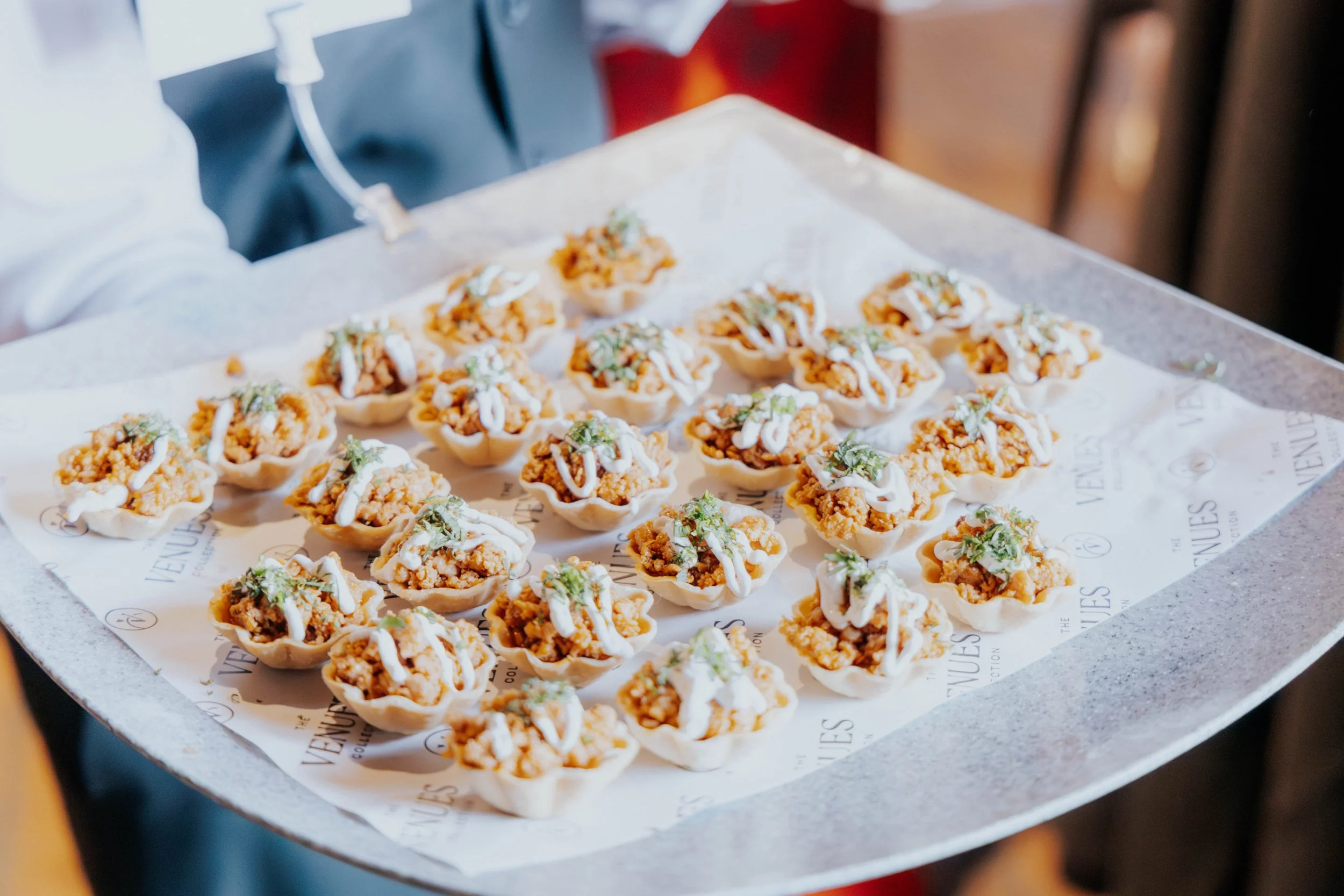 Tray of bite-sized appetizers in pastry shells topped with white sauce and herbs on branded paper liner.