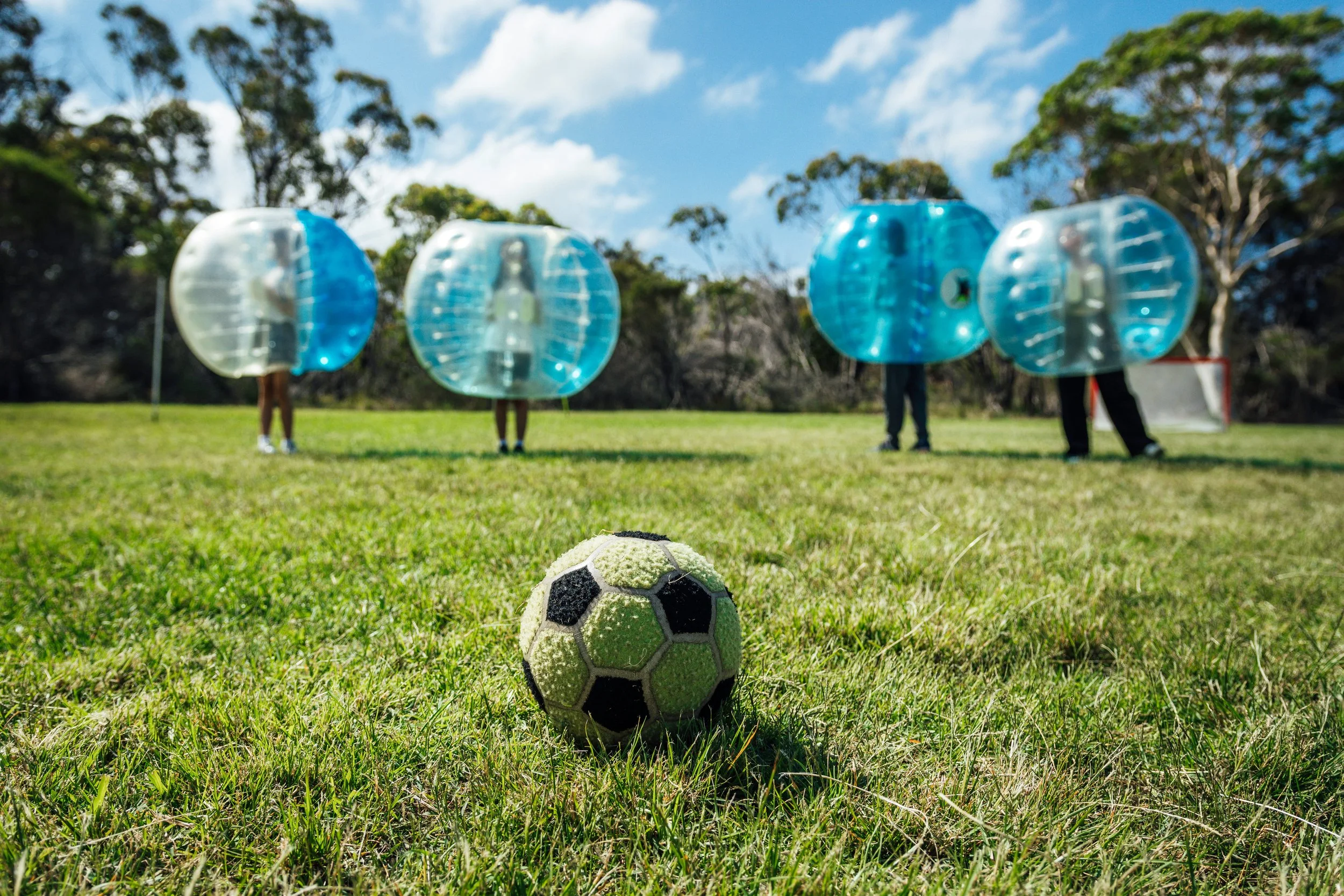 Soccer ball on grass field with four people in bubble suits in the background beneath a blue sky with clouds.