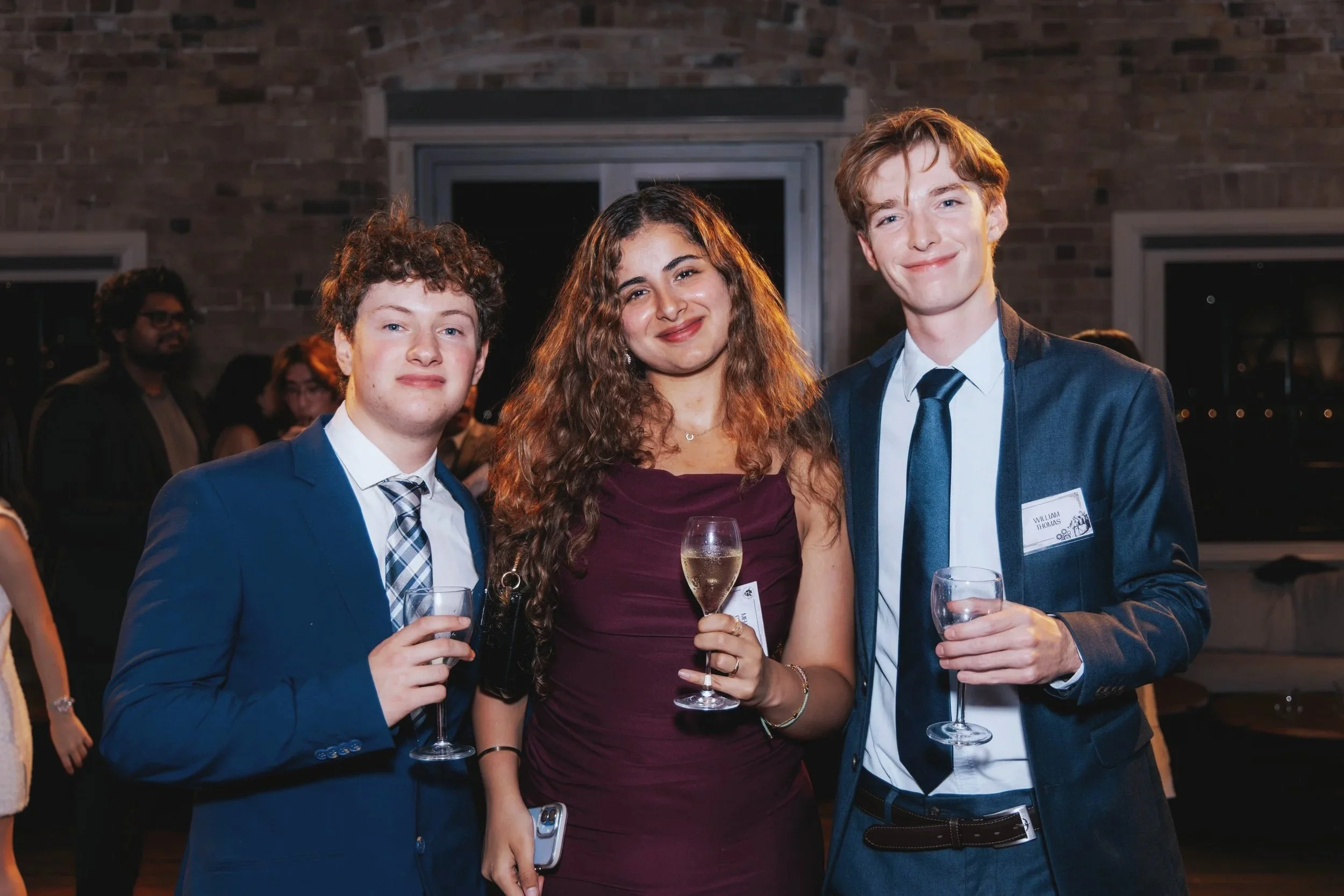 Three young adults at a semi-formal event holding glasses of champagne, smiling in front of a brick wall with other people in the background.