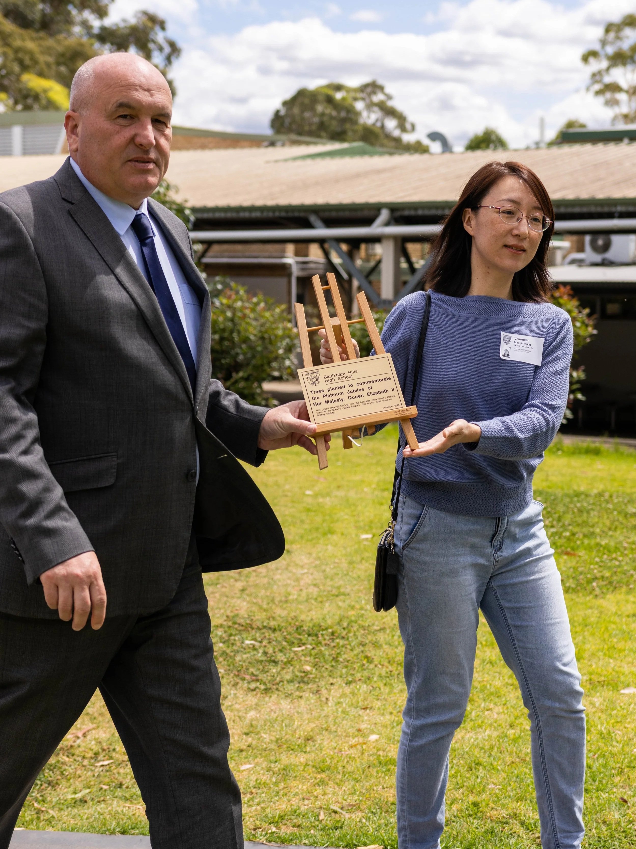A man and a woman are outdoors in a grassy area, holding a small wooden monument with a plaque. The man is in a dark suit and tie, and the woman is wearing glasses, a blue sweater, and jeans. They appear to be participating in a memorial or commemorative event.
