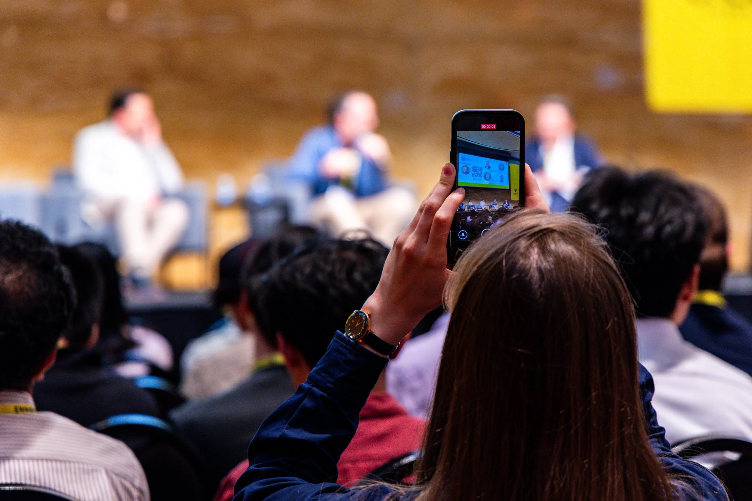 Person recording a panel discussion at a conference with a smartphone.