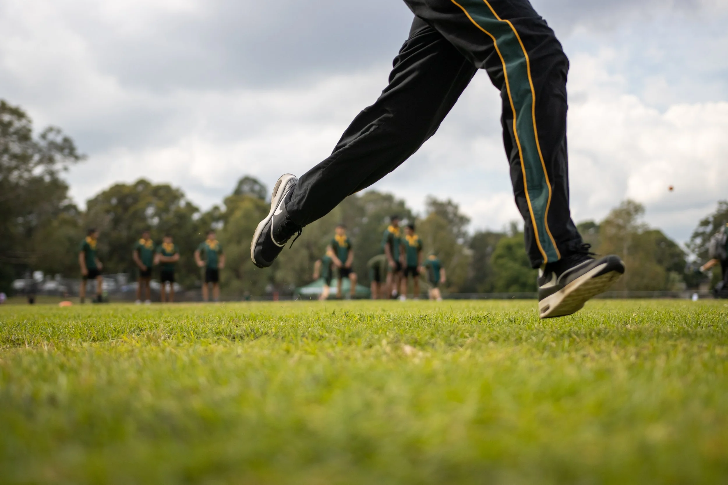 Person in black athletic pants and sneakers running or jumping on a grassy field during a sports practice or game with a group of people in the background.