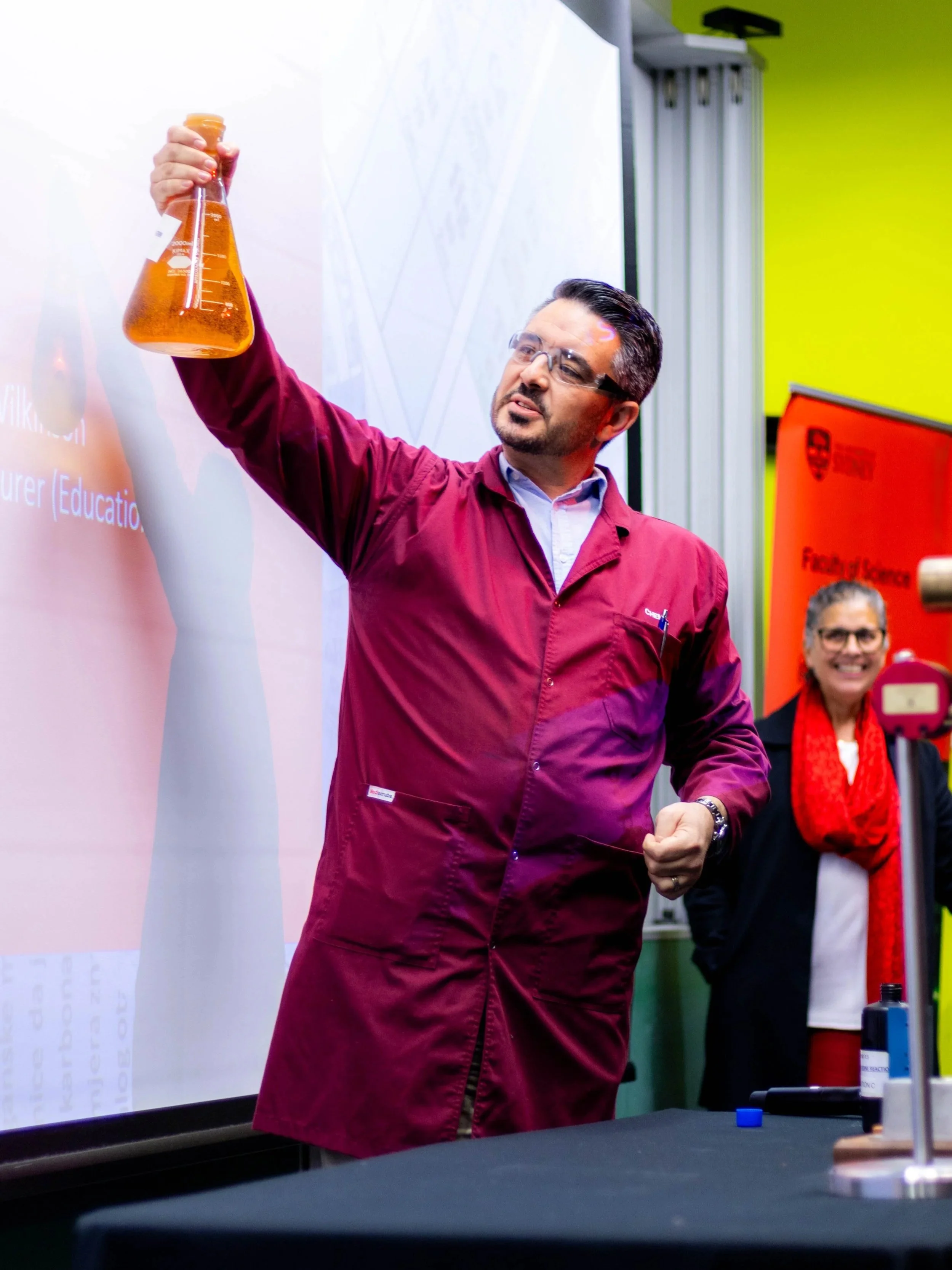 Man in a maroon lab coat demonstrating a chemical experiment with an orange liquid in a laboratory or classroom setting, with a woman smiling in the background.