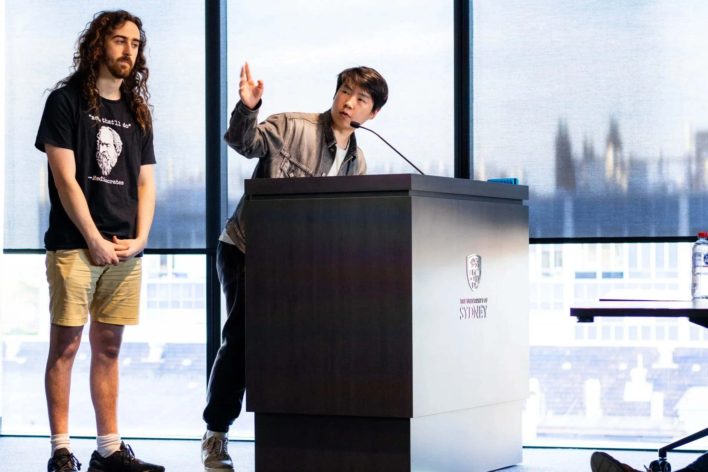 Two young men at a university lecture or presentation: one speaking at a podium with a university emblem, and the other standing nearby. The background features large windows with cityscape views.