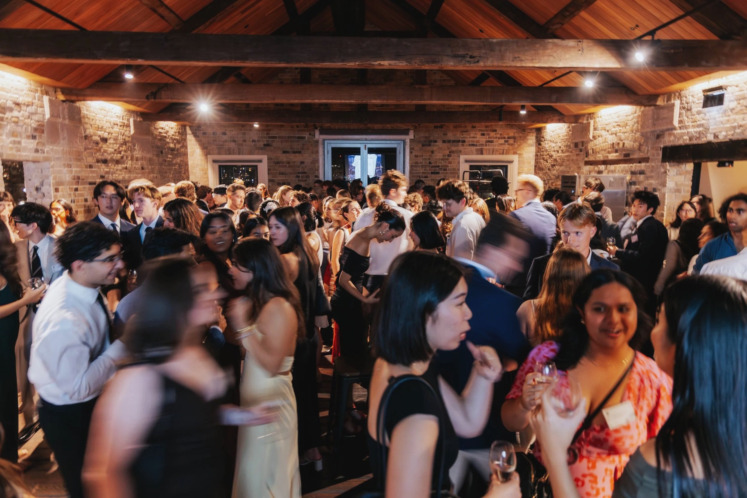 A crowded party or social gathering in a room with brick walls and wooden ceiling beams, featuring diverse people dressed in formal and semi-formal attire, some holding glasses of wine, engaging in conversation and socializing.