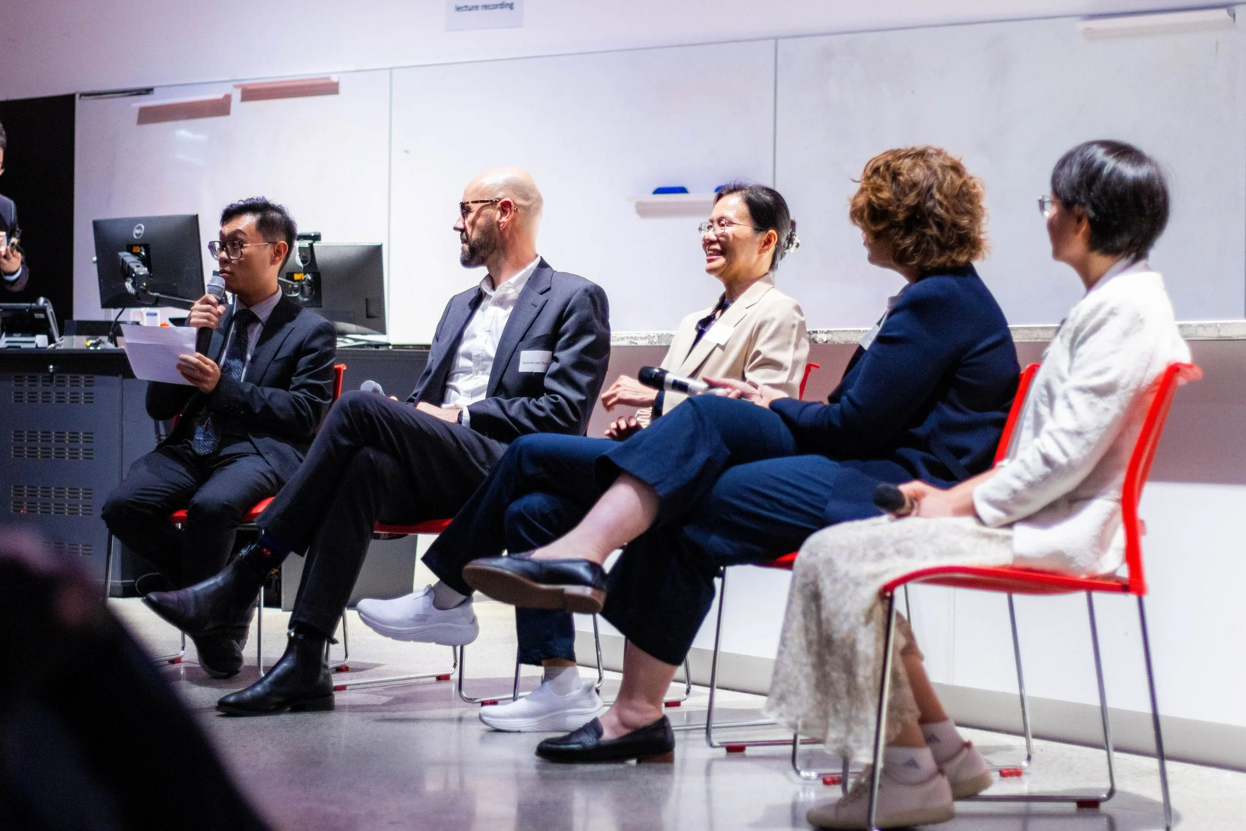 Panel of five diverse individuals seated on stage during a discussion, with one person speaking into a microphone and a whiteboard in the background.