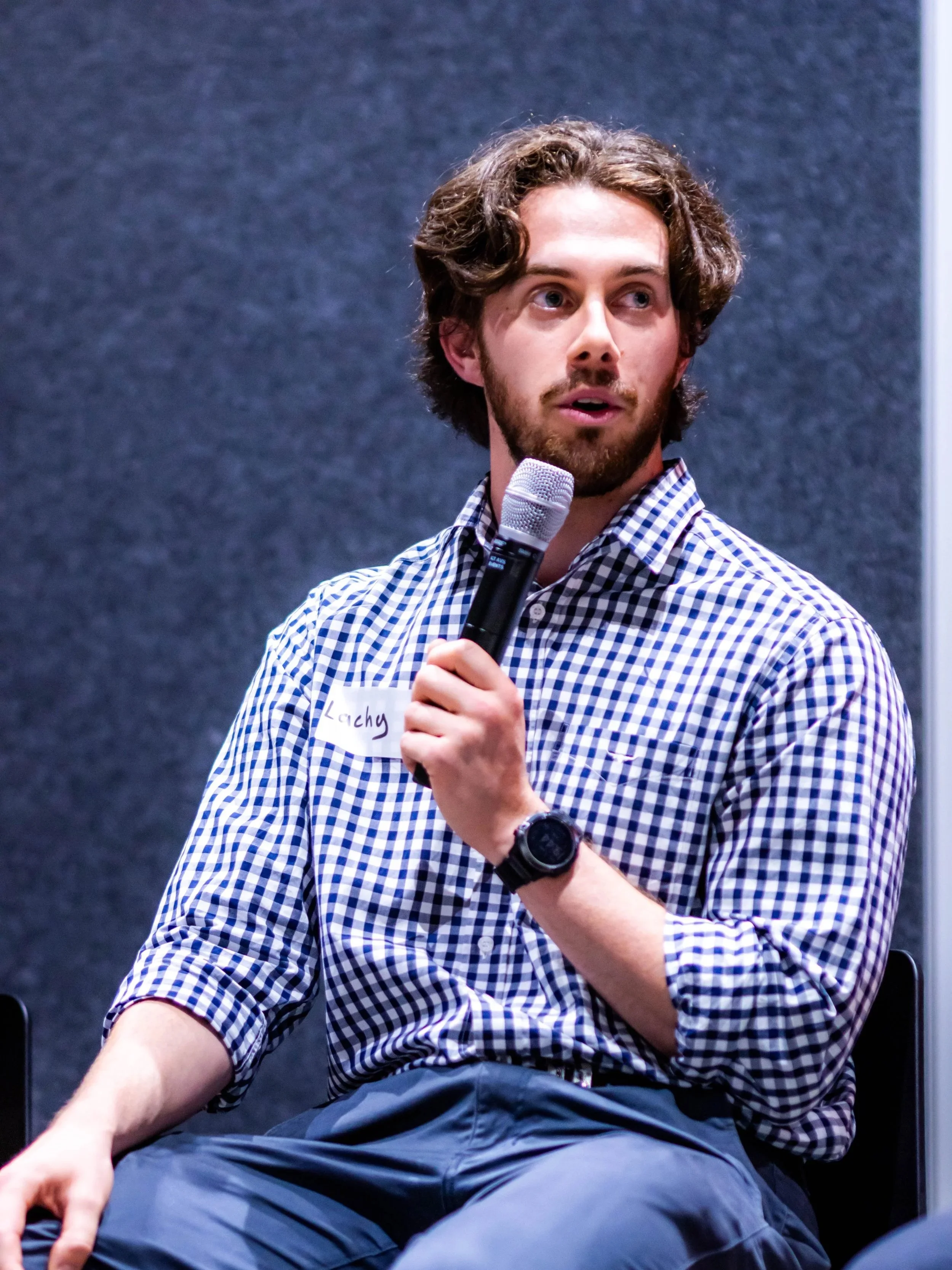 Young man with curly hair and beard sitting, holding a microphone, wearing a checkered shirt, with a name tag that says "Lachy," and a black watch on his left wrist, in front of a gray background.
