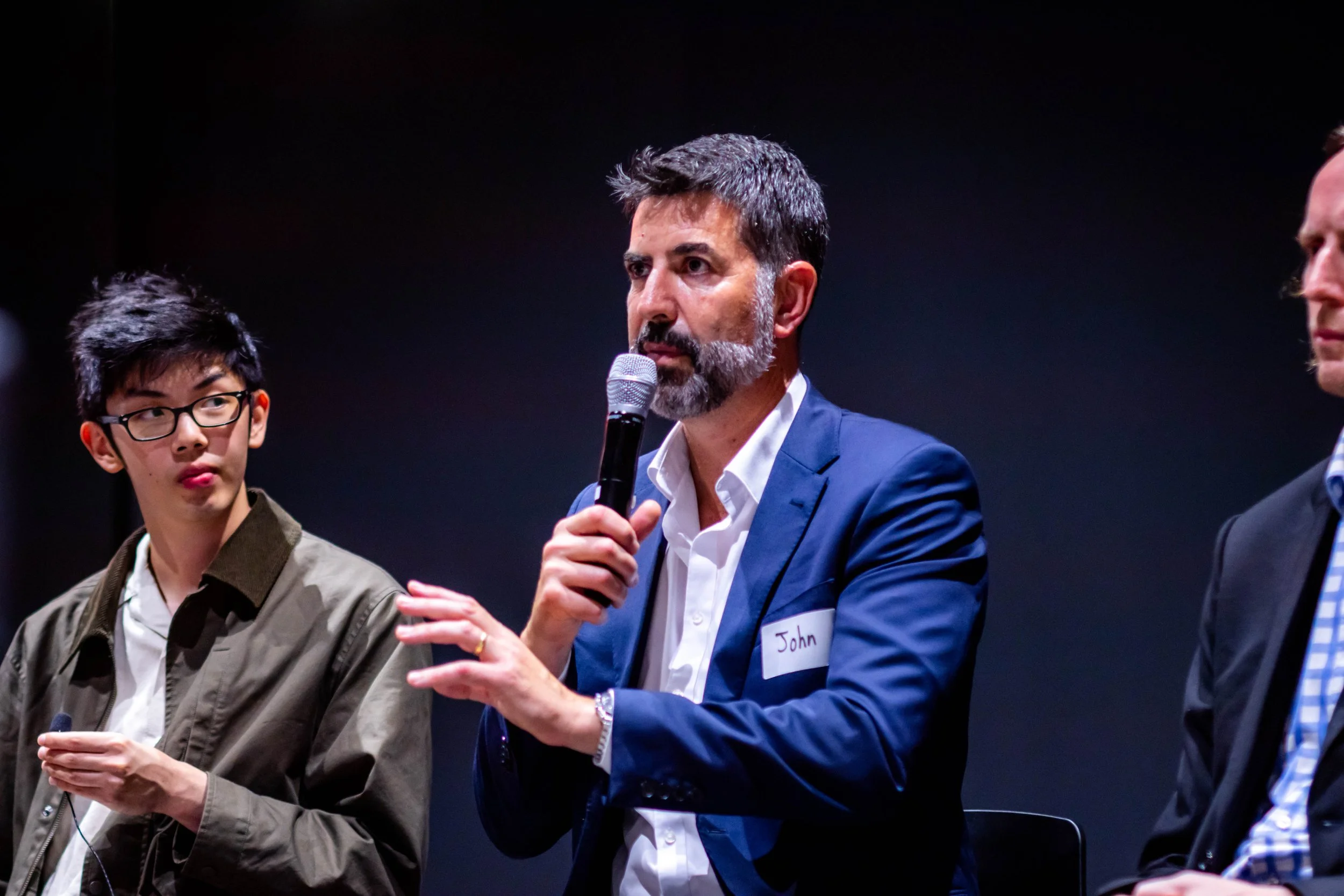 Three men sitting on a stage, one in the middle holding a microphone and speaking, wearing a suit with a name tag that reads "John." The man on the left has glasses, dark hair, and is dressed in a jacket. The man on the right, partially visible, is wearing a suit with a striped tie.