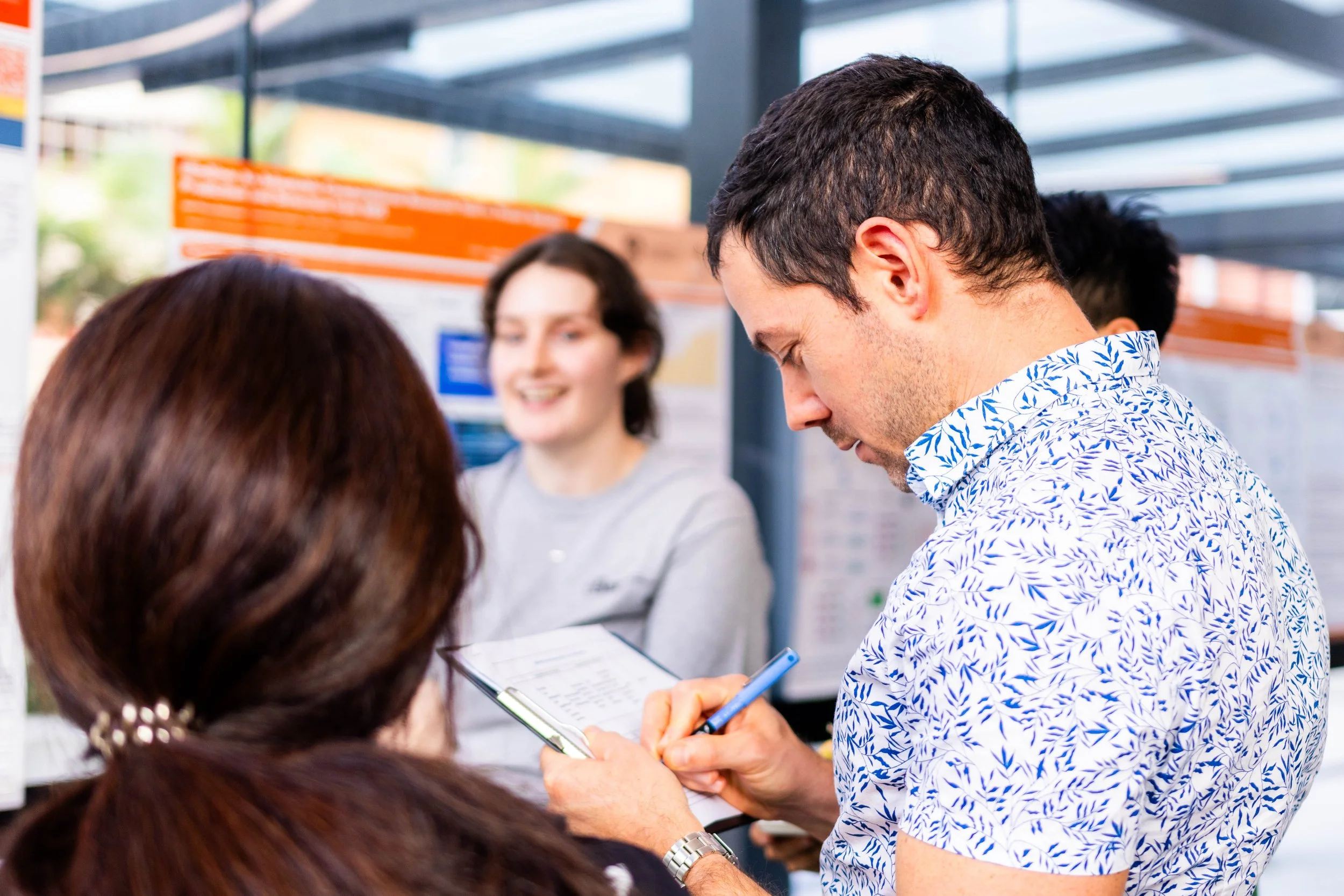 A man looks down at a device and interacts with a touchscreen device at an airport check-in counter, with other travelers around him and a smiling woman in the background.