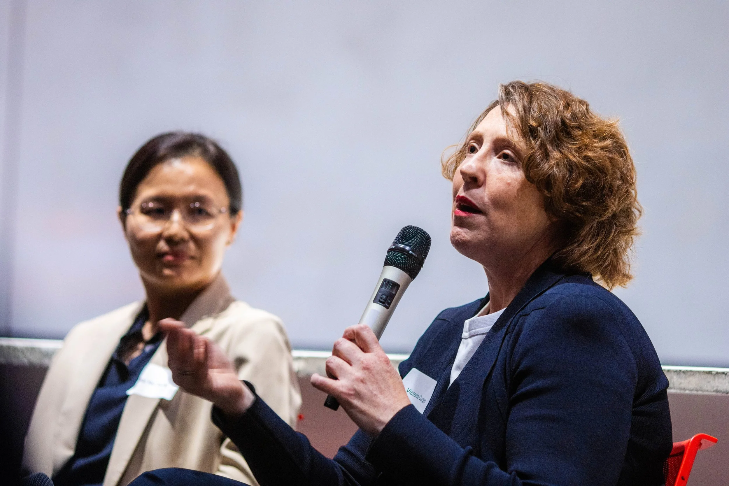 Two women engaging in a discussion, one holding a microphone and speaking, the other listening with a slight smile, against a plain background.