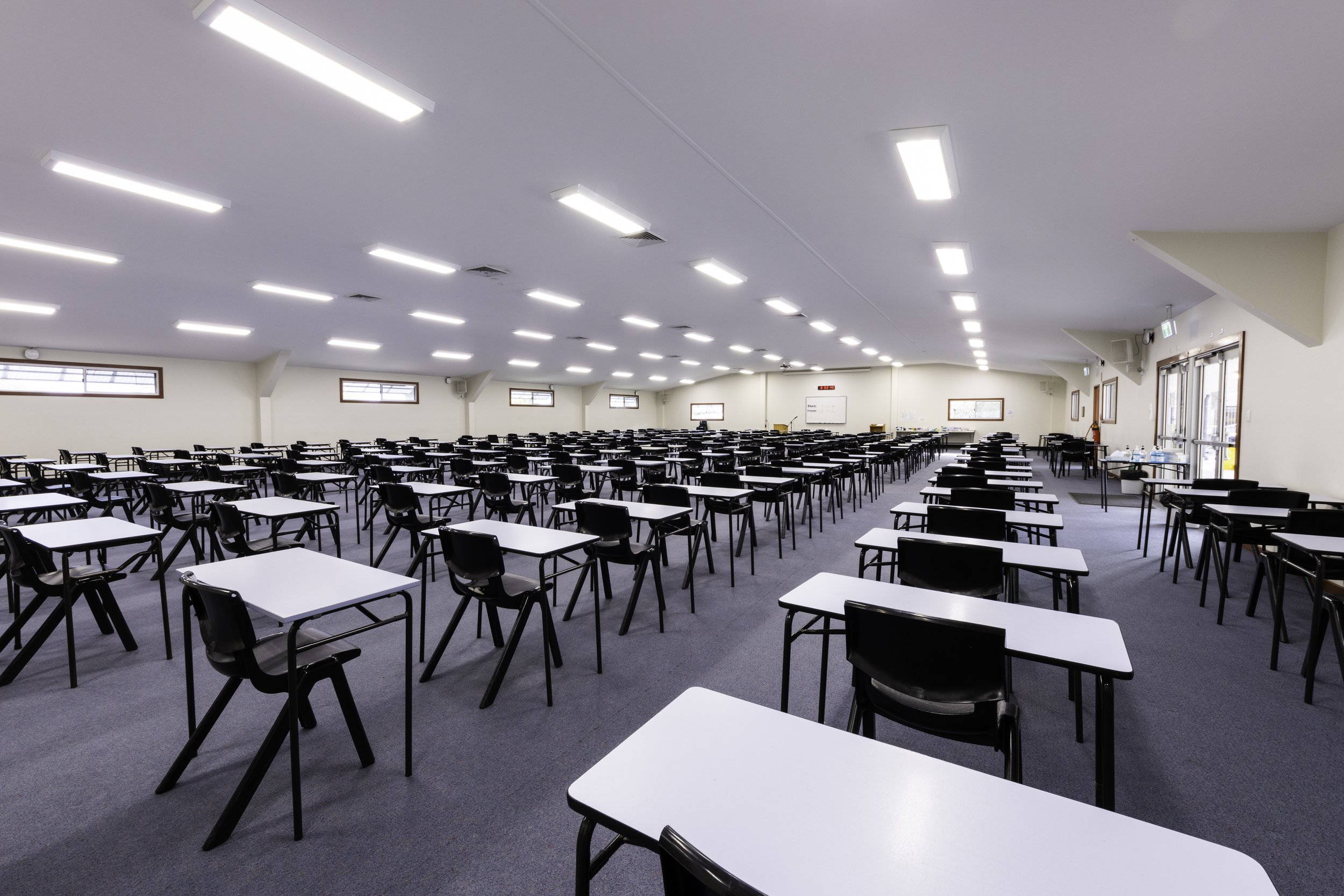 An empty classroom or examination hall with rows of white desks and black chairs, bright ceiling lights, and windows on the side walls.