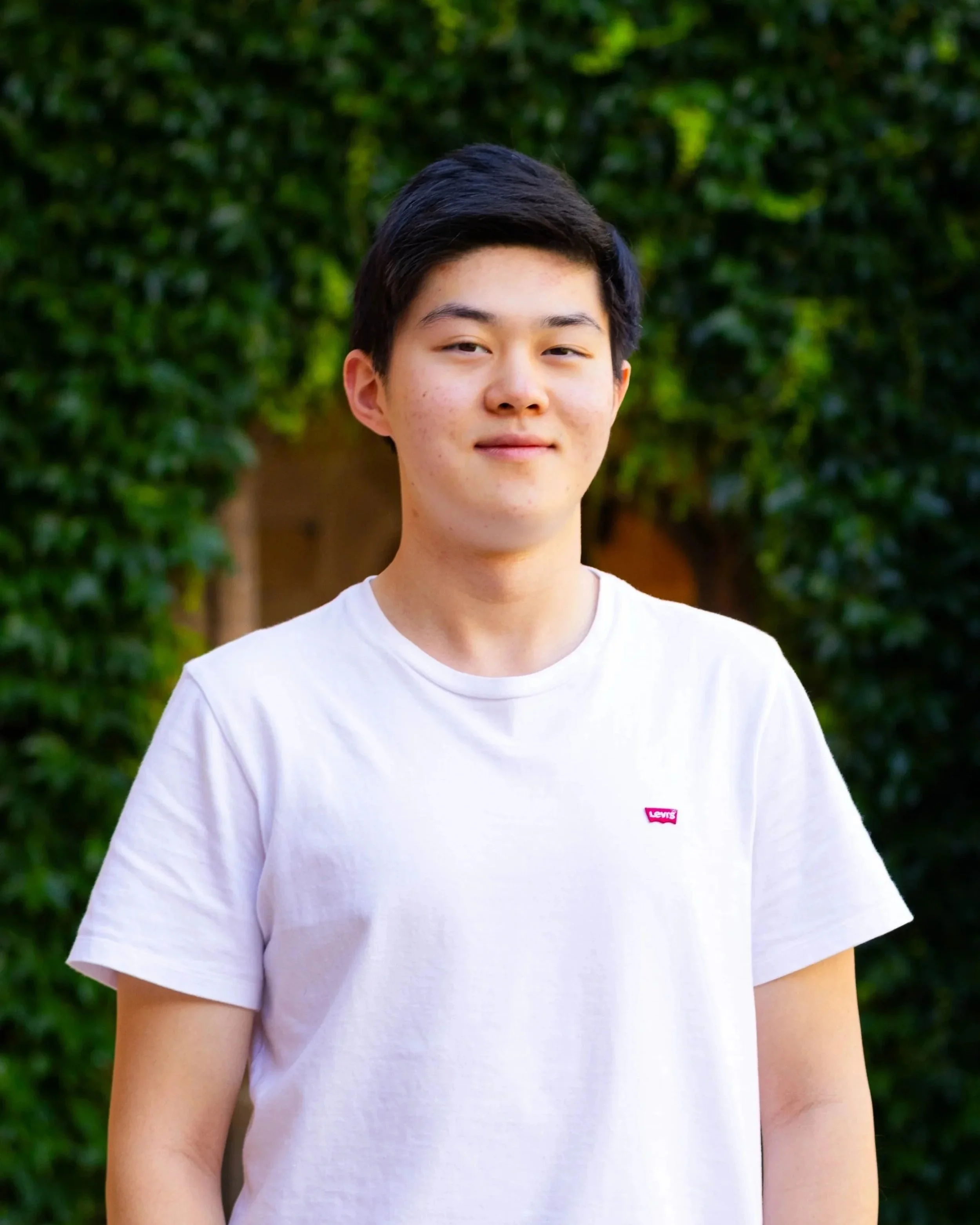 A young man with dark hair, wearing a white Levi's t-shirt, standing outdoors in front of a green hedge, looking at the camera with a slight smile.