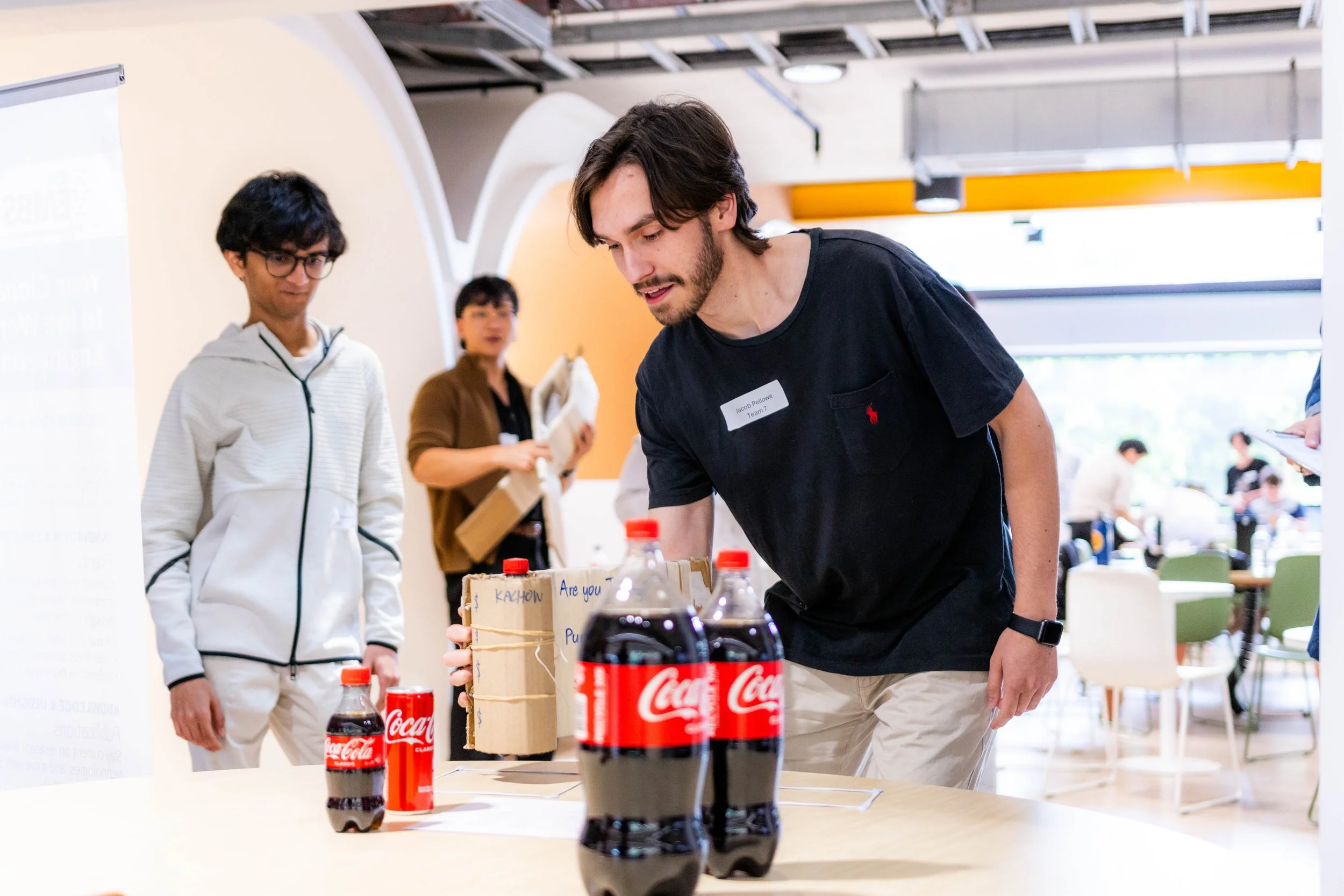 Man arranging Coca-Cola bottles on a table at an indoor event, with other people in the background.