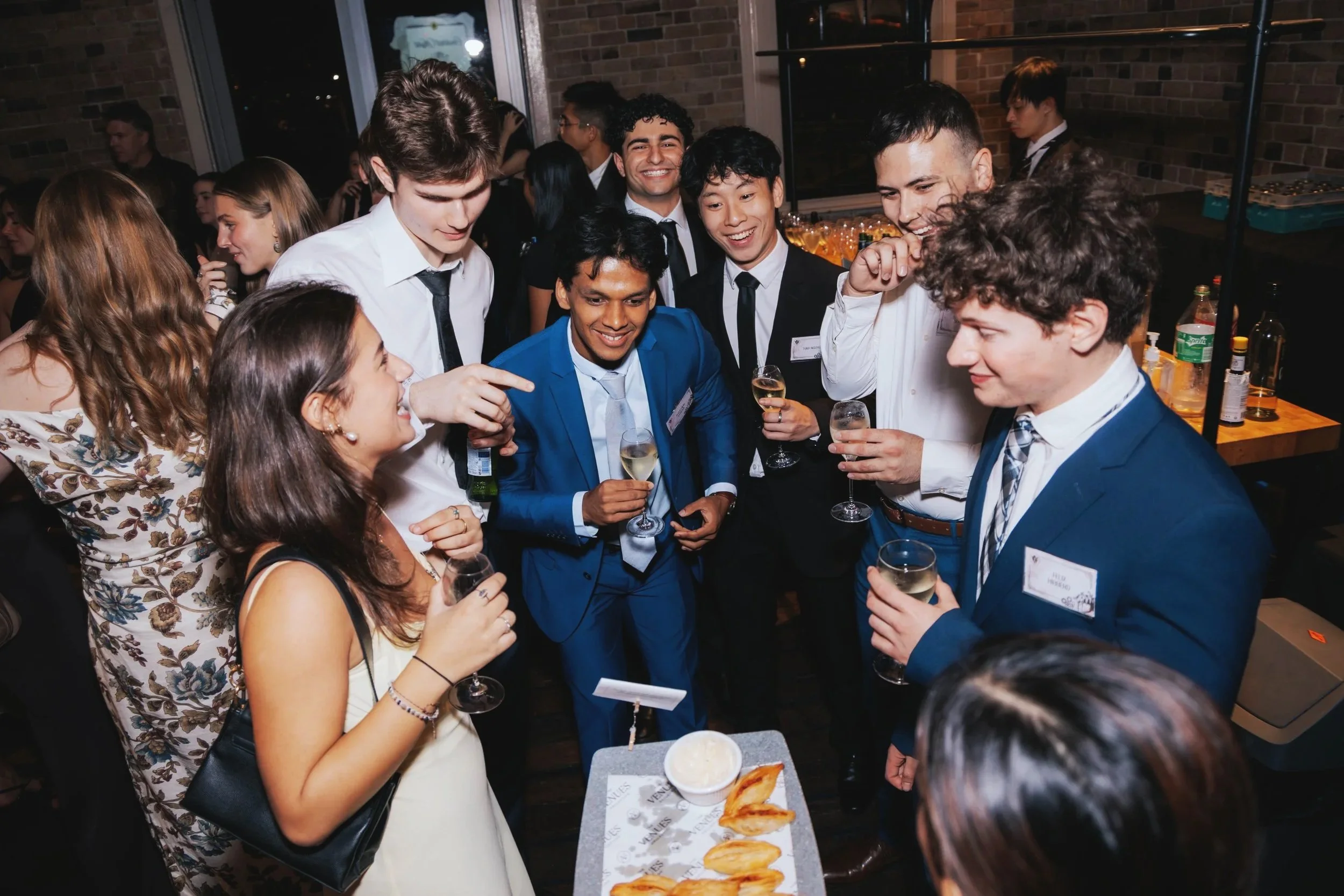 Group of young adults at a social event, holding drinks and engaging in conversation, indoors at a party or celebration.