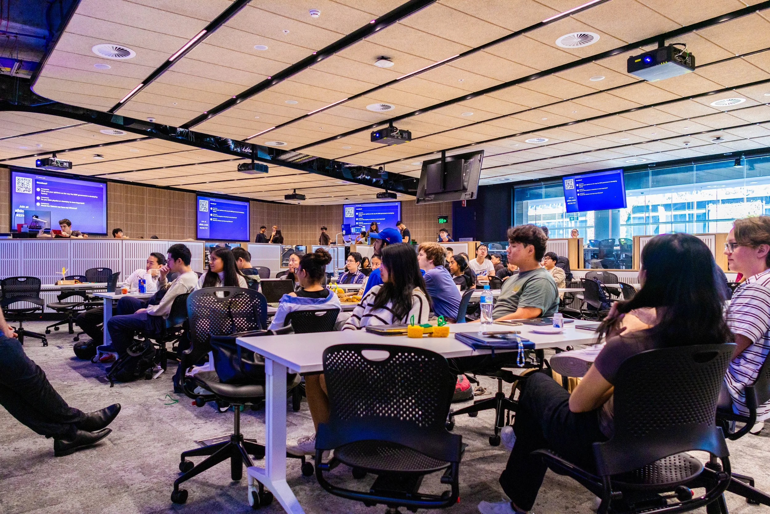 A large conference room filled with people seated at tables, attending a presentation on multiple large screens at the front of the room. The room has a modern design with a high ceiling and large windows.
