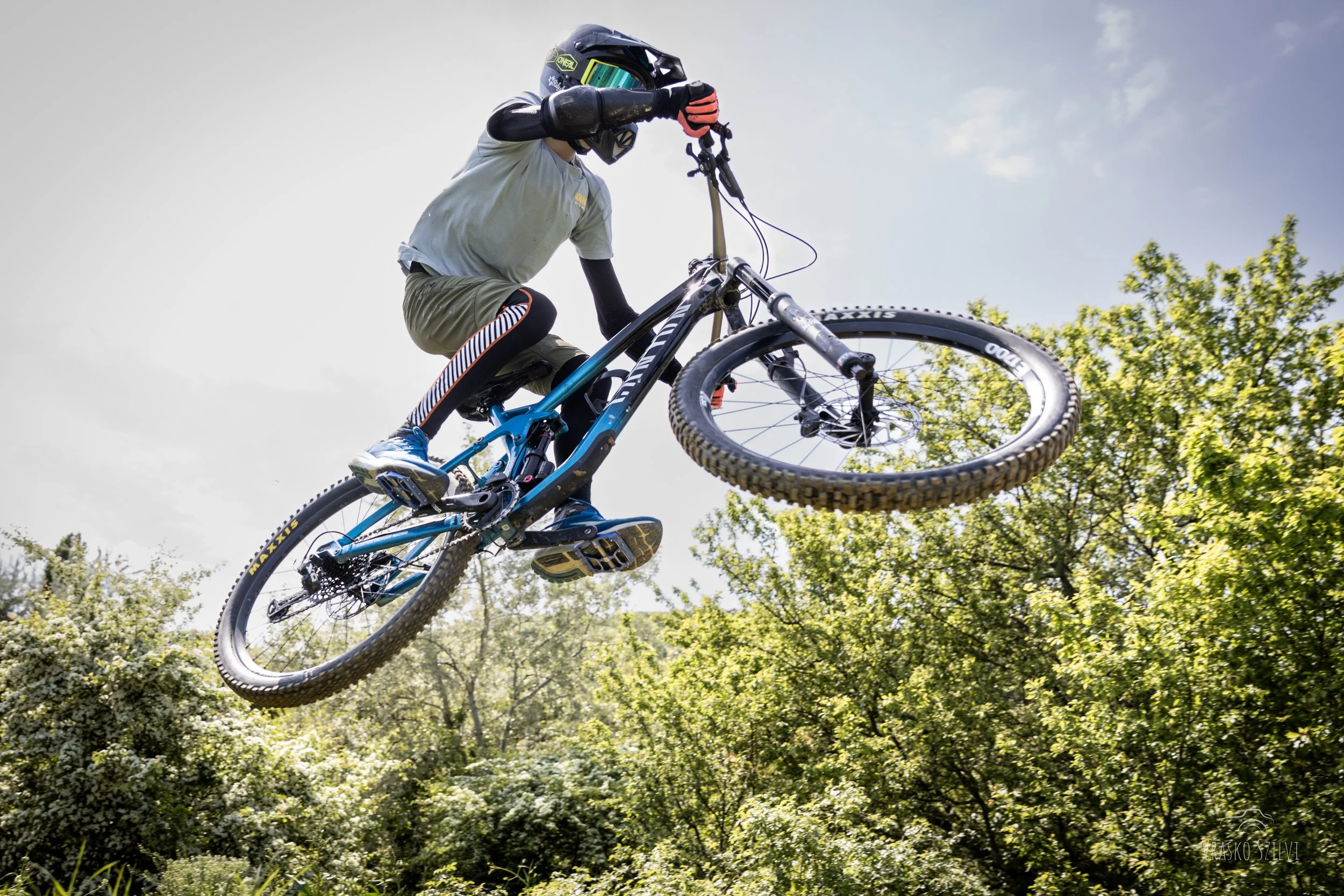 A mountain biker wearing a helmet and riding gear jumping off a ramp over a lush green landscape.