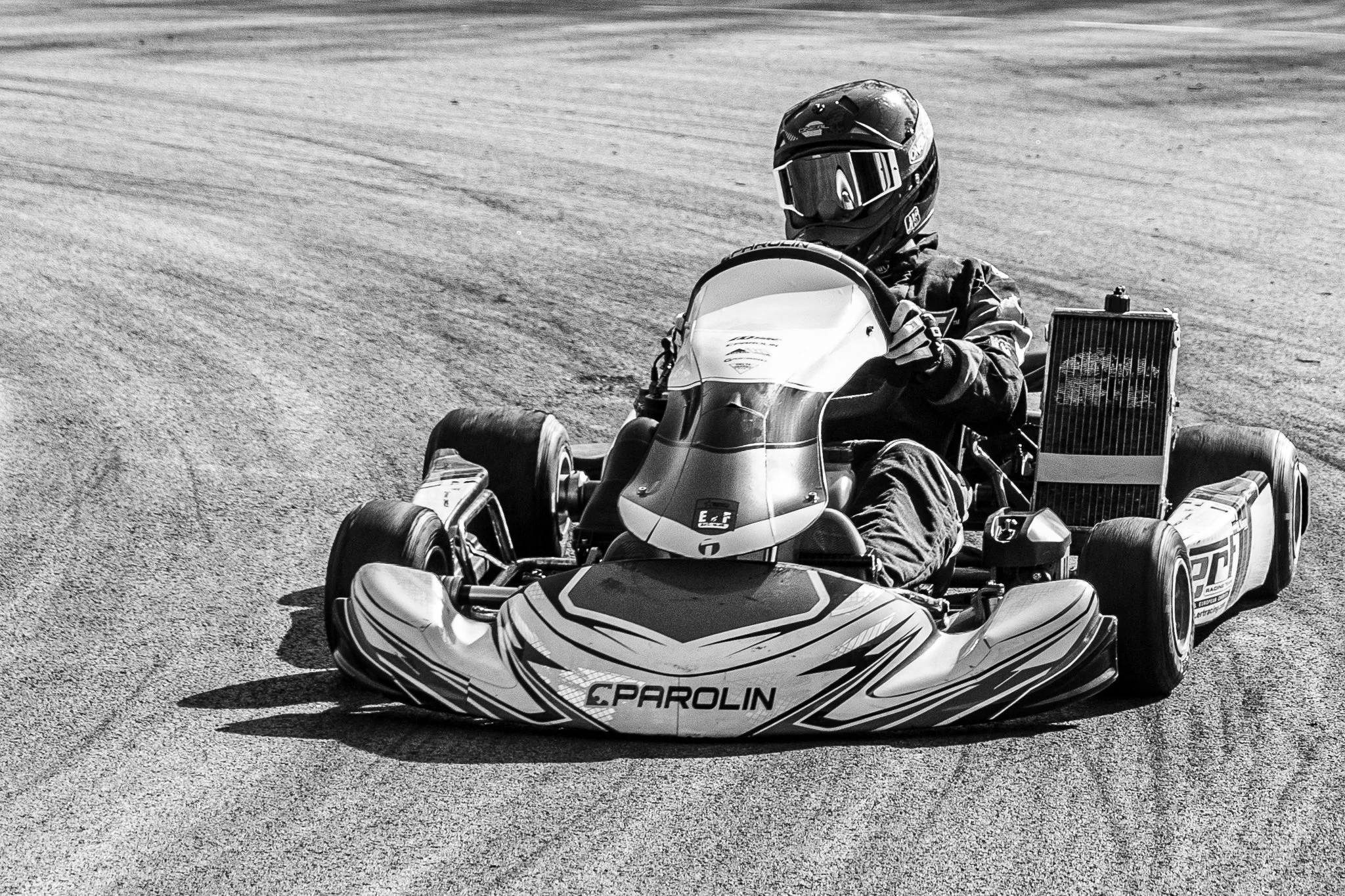 A person driving a go-kart on a paved track, wearing a helmet and racing suit, with the front of the kart labeled "Parolin."