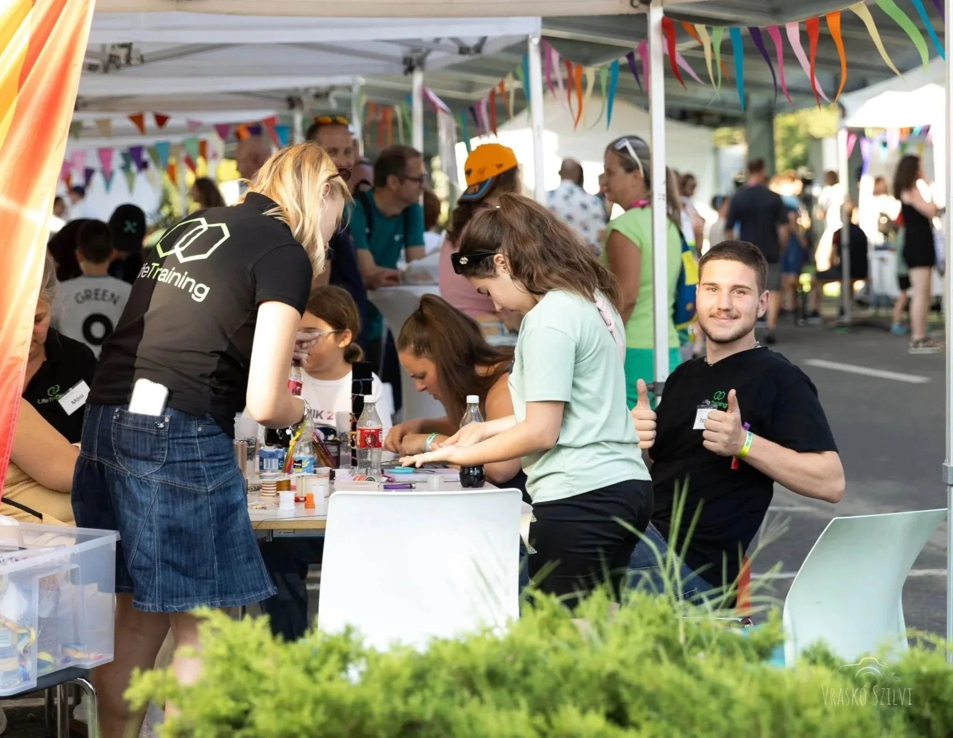 People at an outdoor festival or fair participating in craft activities under colorful bunting decorations. A young man giving a thumbs-up and smiling, seated to the right.