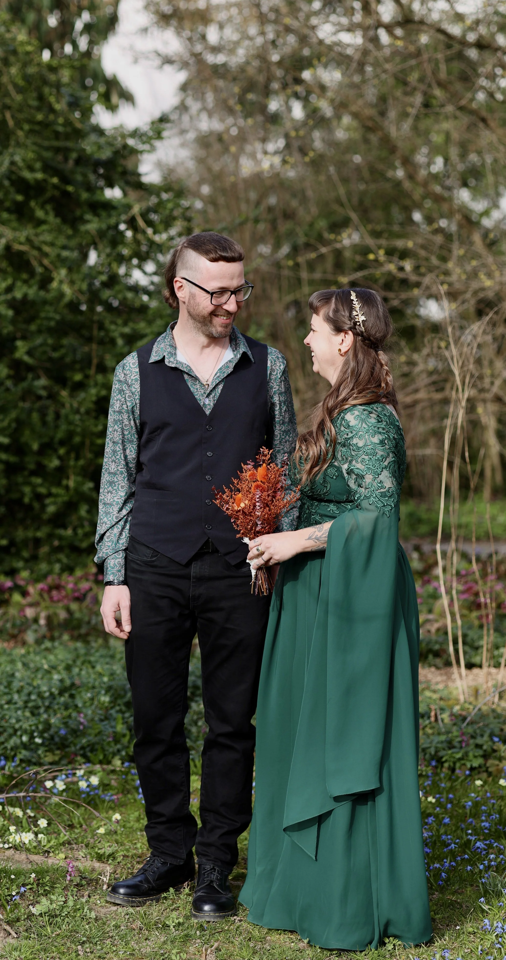 Ein Mann und eine Frau stehen in einem Garten. Der Mann trägt eine Brille, ein Hemd mit Muster und eine schwarze Weste, während die Frau ein elegantes grünes Kleid mit Stickereien trägt und eine Blume im Haar hat. Sie hält einen Blumenstrauß in den H