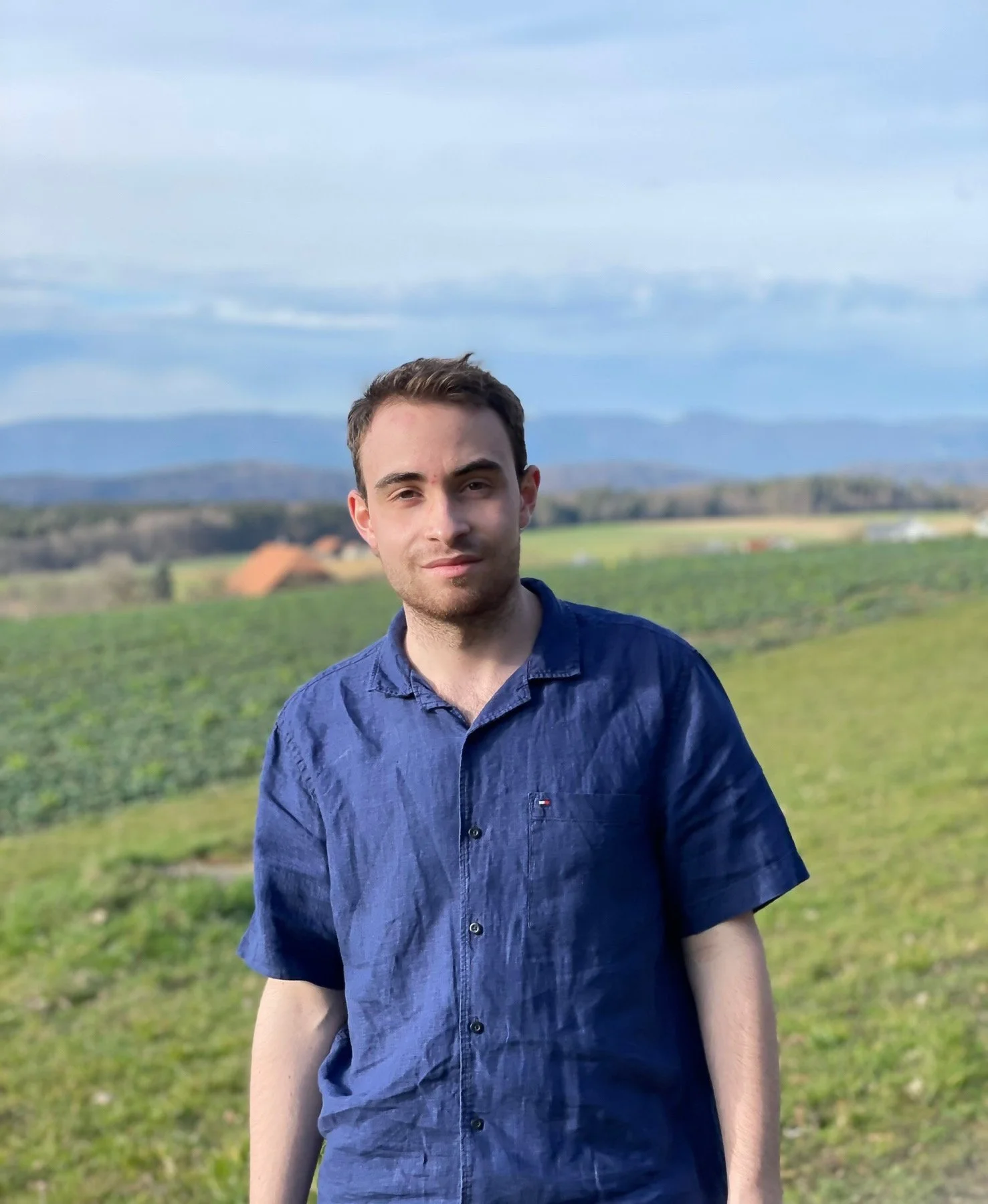 Ein junger Mann trägt ein blaues Hemd und steht vor einer ländlichen Landschaft mit Feldern, Häusern und Bergen im Hintergrund, unter einem bewölkten Himmel.