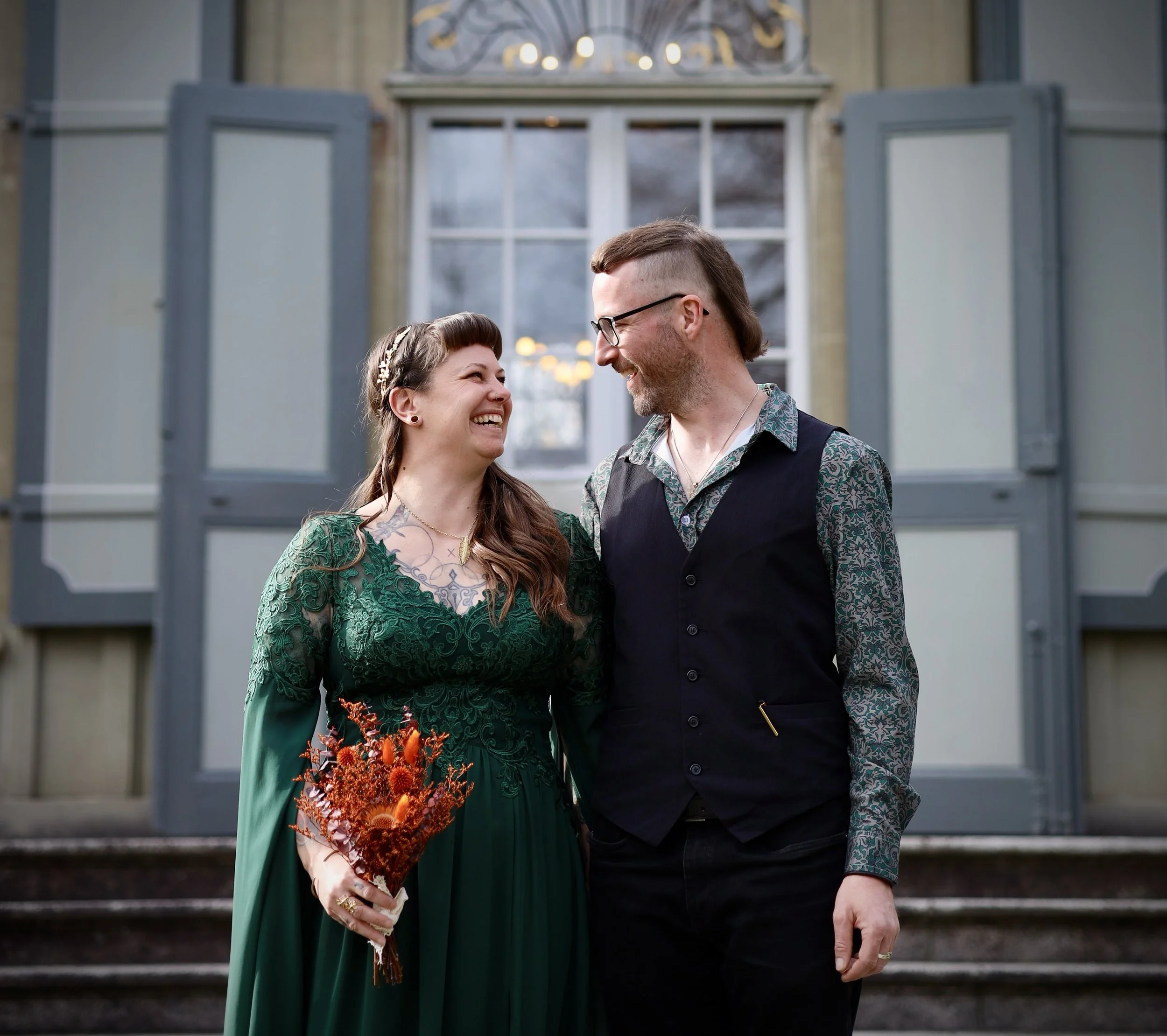 Ein verliebtes Paar steht vor einem Haus, lächelt sich an und hält Hände. Die Frau trägt ein grünes Kleid mit Spitzen und hält einen Blumenstrauß, der hauptsächlich orangefarbene Blumen enthält. Der Mann trägt eine Brille, ein gewaschenes Hemd und ei