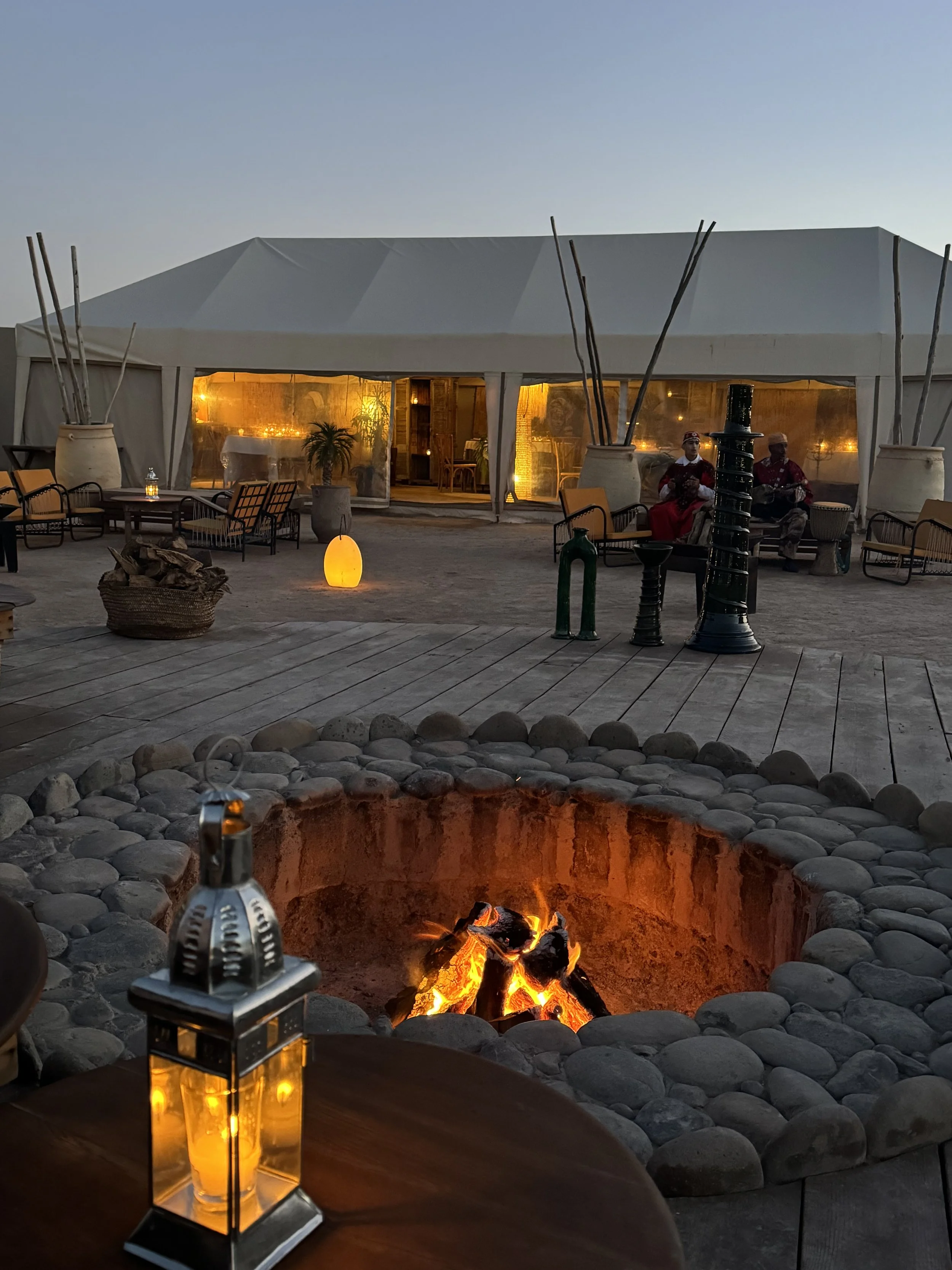 Ein Zelt im Abendlicht, im Inneren warm erleuchtet mit Pflanzen und Tischen, im Außenbereich Sitzmöbel. Im Vordergrund ein Feuergrube mit brennendem Holz und Steinen, umgeben von Steinen. Es stehen Steine, Lampen und Dekorationen im Garten.