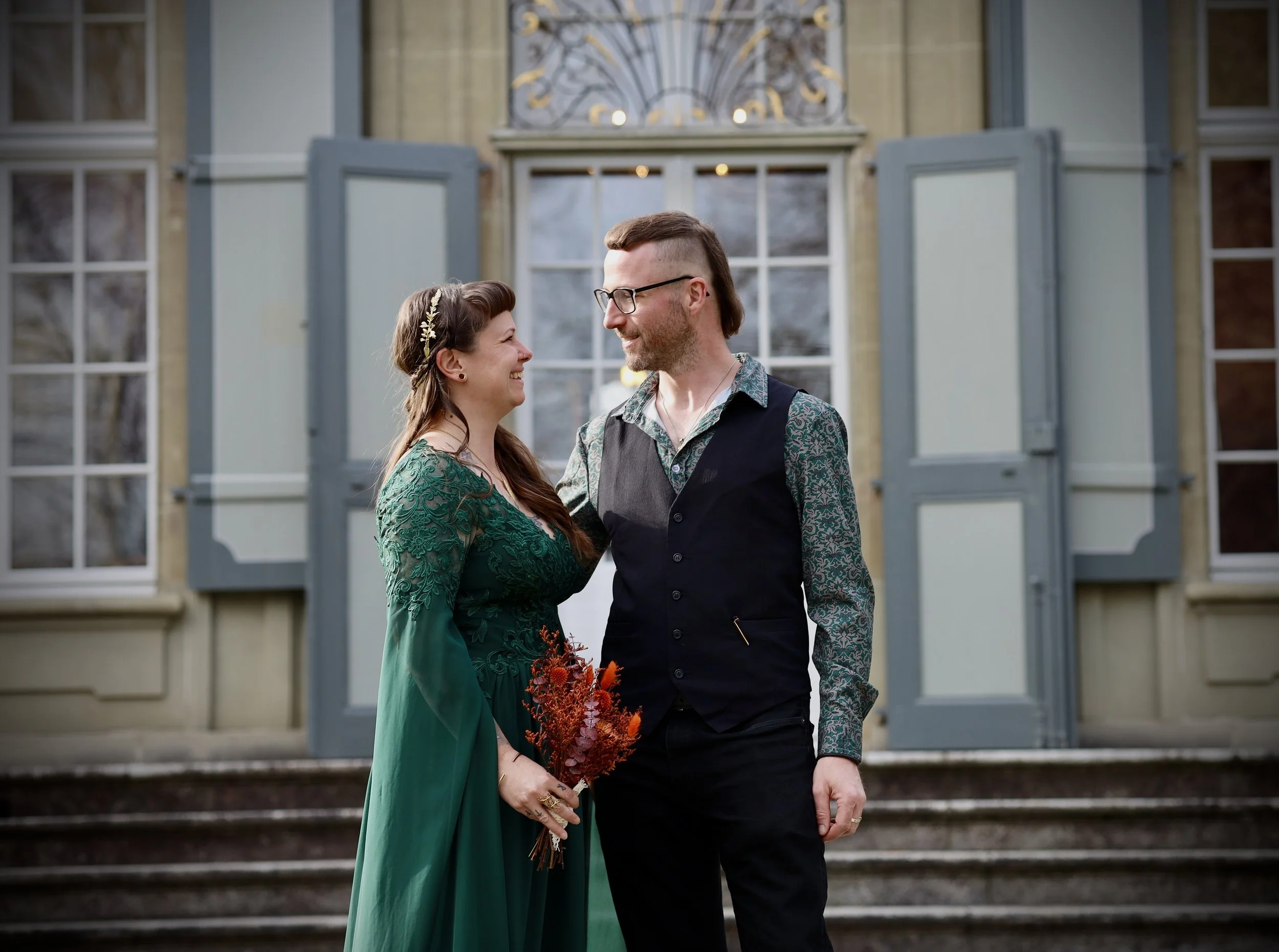 Ein Paar stehen vor einem alten Haus, lächeln sich an und halten sich an den Händen. Das Mädchen trägt ein elegantes grünes Kleid und eine goldene Haarkranz, der Mann trägt eine geblümte Bluse und eine Weste. Das Mädchen hält einen Blumenstrauß in de