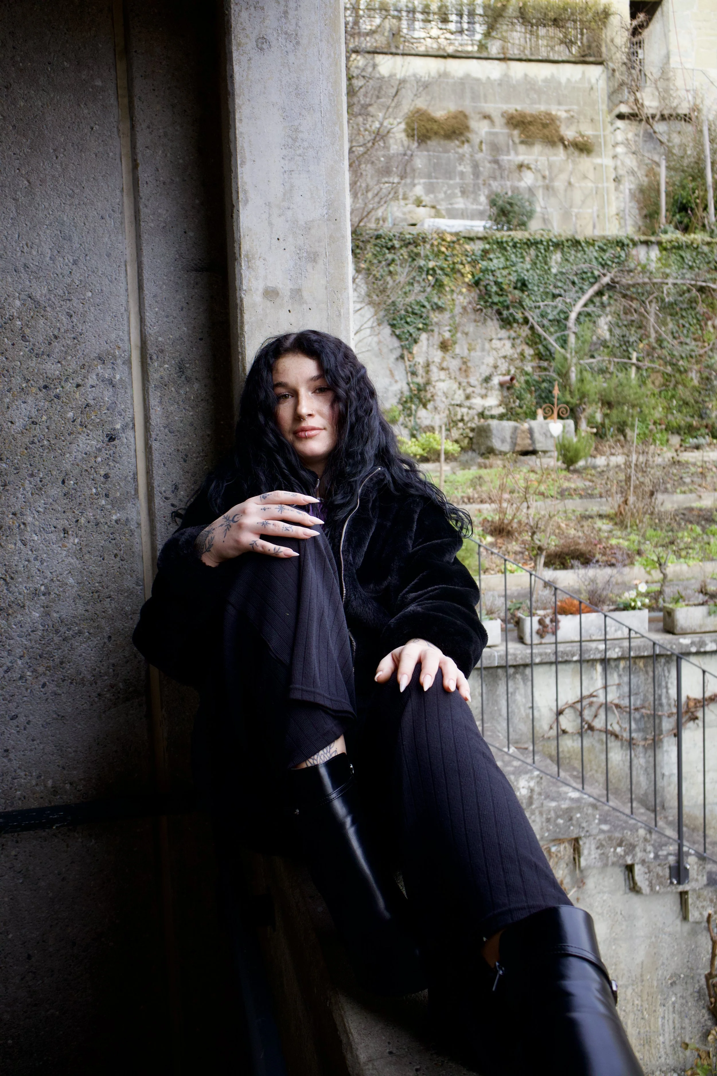 Junge Frau mit schwarzer Lockenfrisur und Tattoos, sitzt entspannt an einer Betonwand im Garten, trägt eine schwarze Jacke und Stiefel, Blick direkt in die Kamera.
