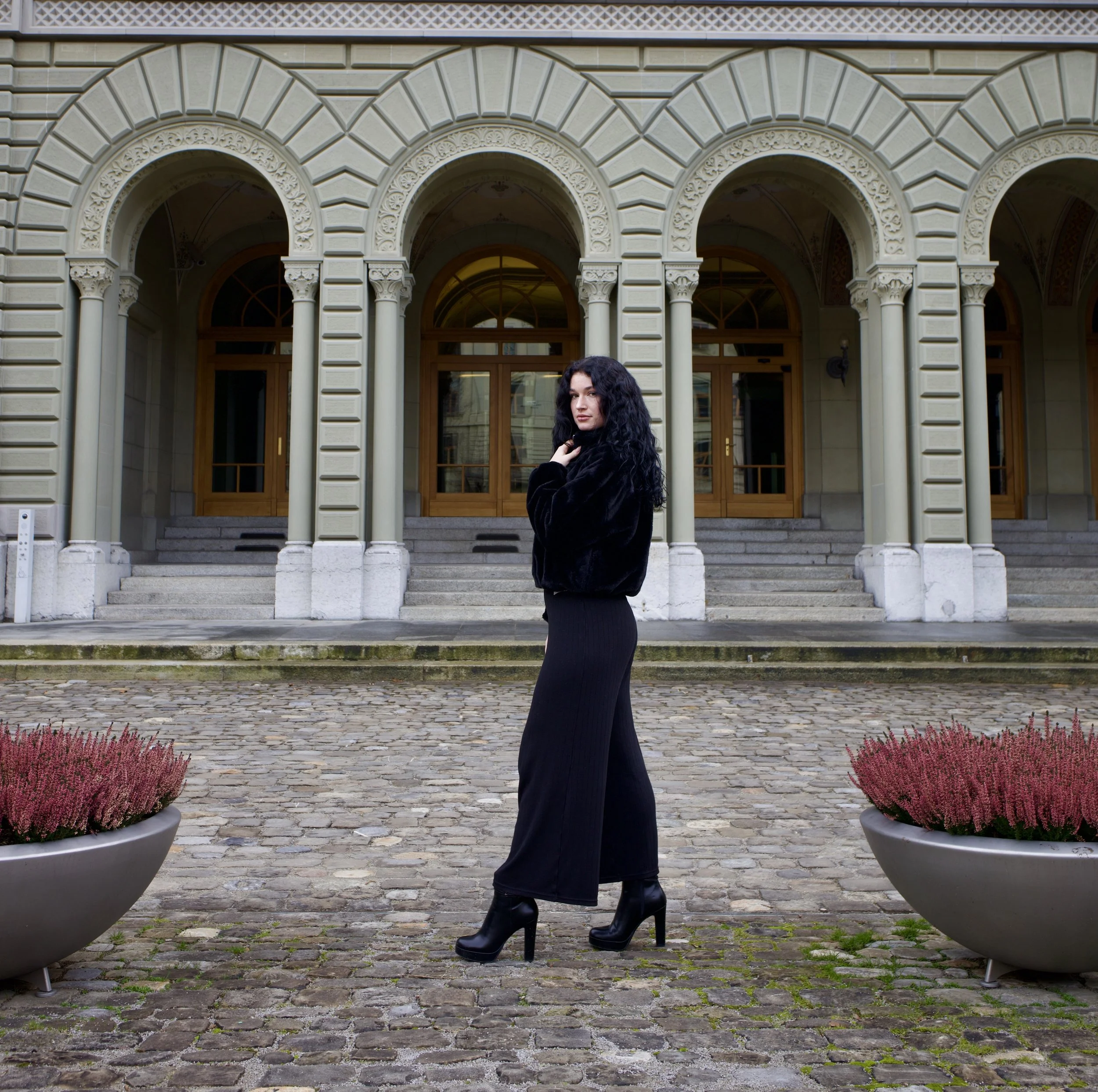 Junge Frau mit schwarzen, lockigen Haaren und dunklem Outfit steht vor historischen Gebäuden mit arched Fenstern und Säulen, im Vordergrund zwei große Pflanzkübel mit pinken Blumen, auf Kopfhöhe.