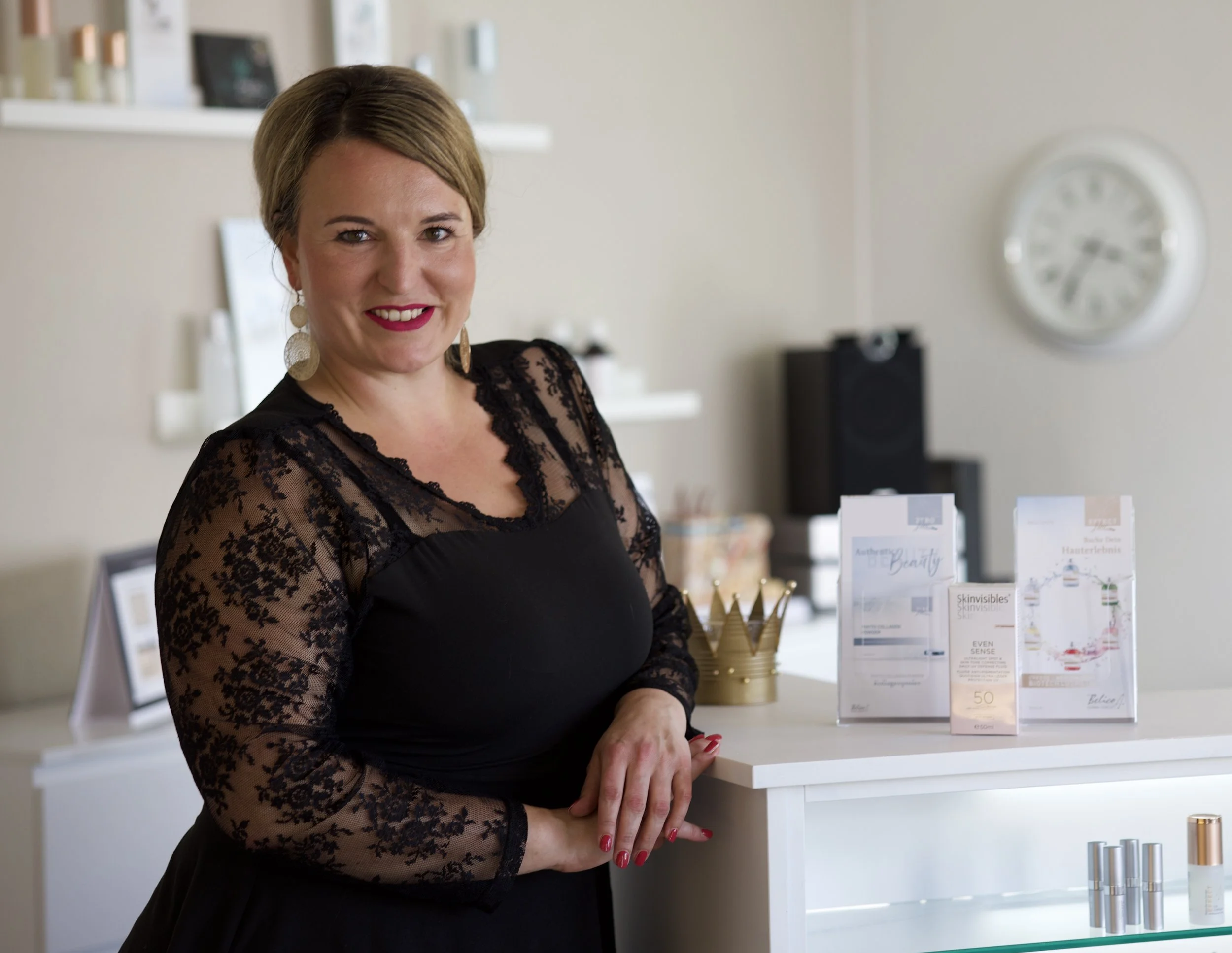 Eine Frau mit einem schwarzen Kleid mit Spitze und großen Ohrringen steht an einem weißen Regal mit Beauty- und Pflegeprodukten. Sie hat braunes Haar, trägt roten Lippenstift und rote Nagellacke.