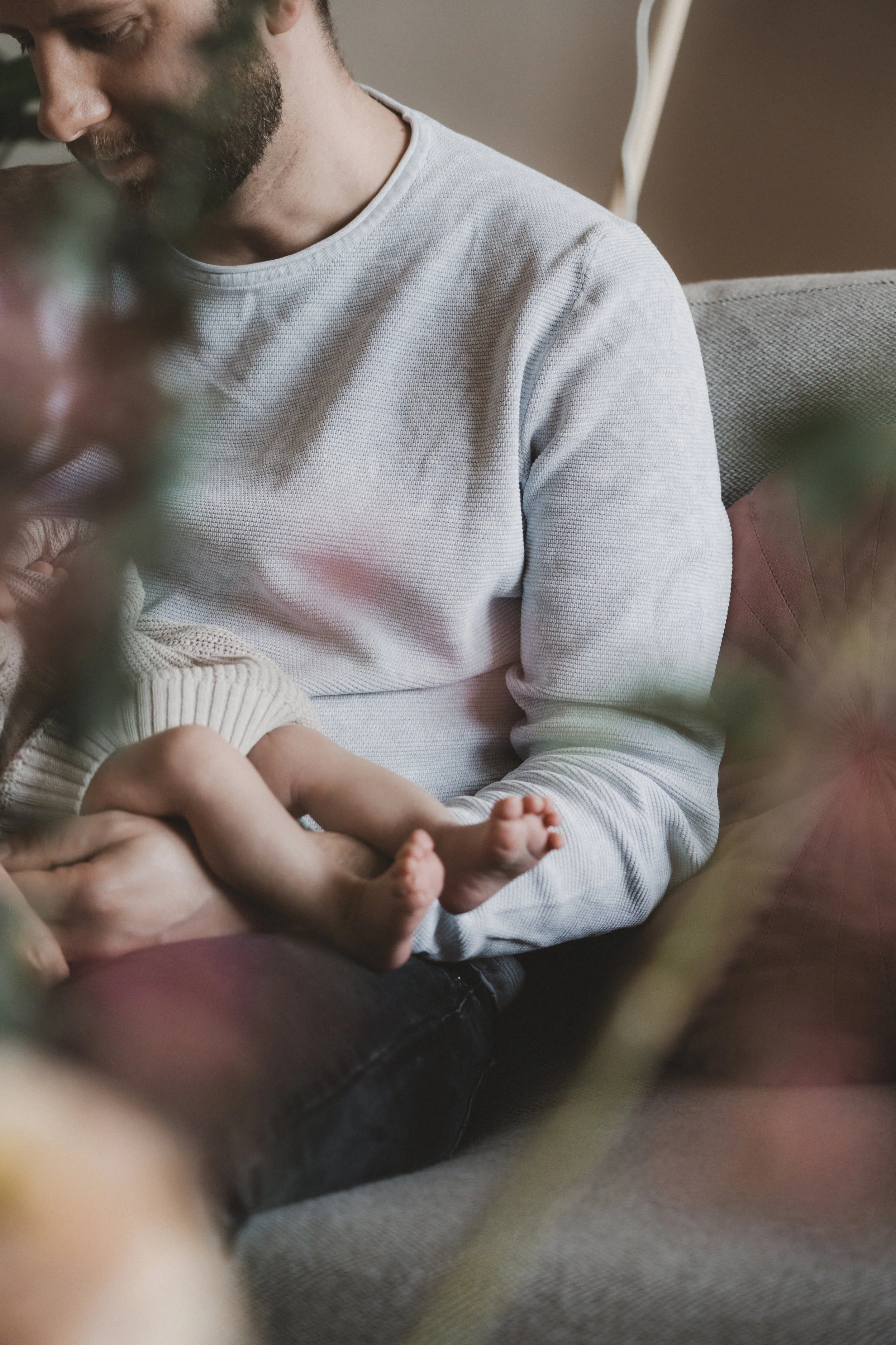Ein Mann hält ein Baby auf seinem Schoß, umgeben von Blumen im Vordergrund, in einem gemütlichen Raum.