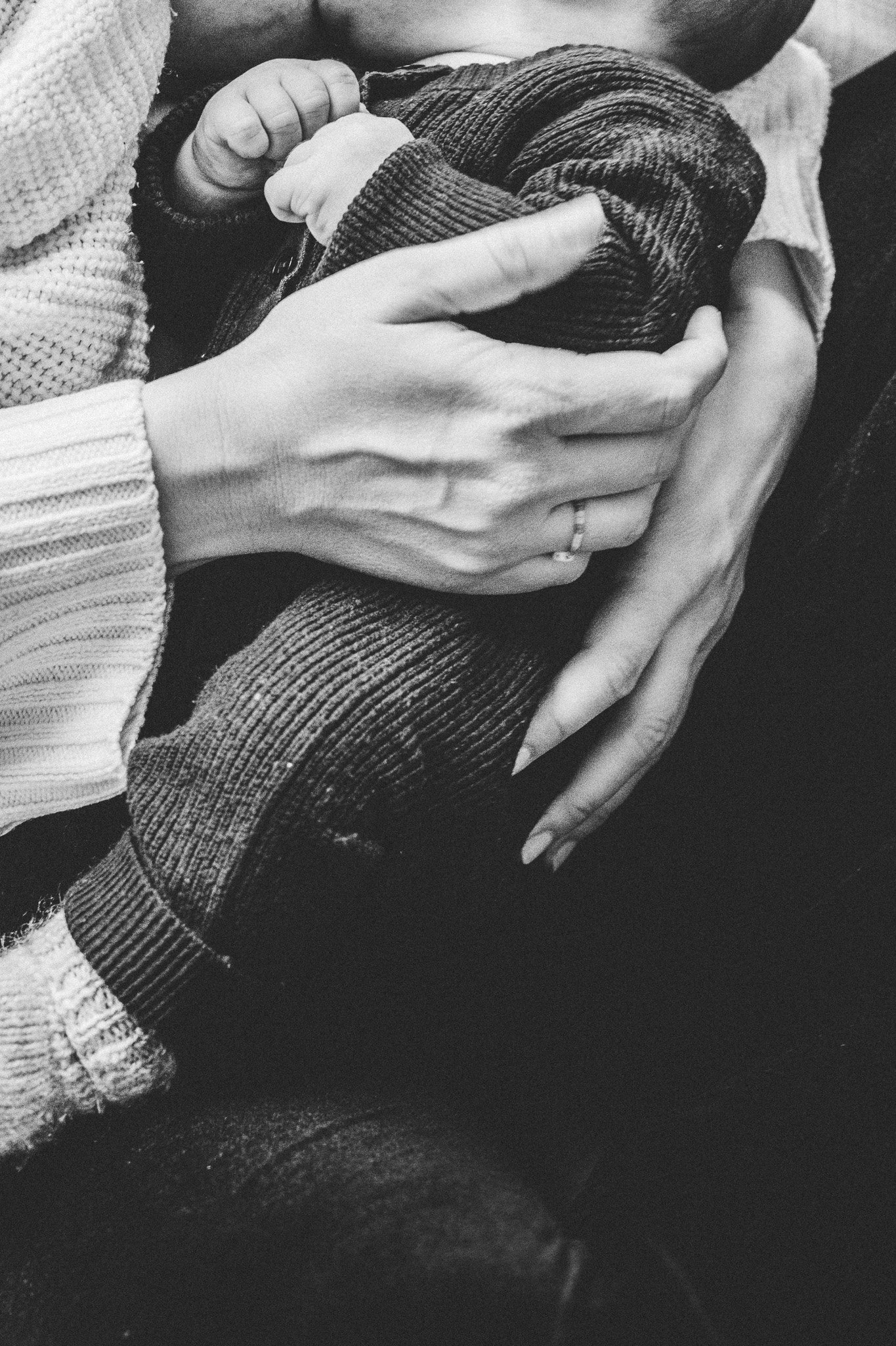 Eine Person hält ein Baby, das in einem Kleid aus Stoff eingewickelt ist. Die Szene ist in Schwarz-Weiß. Alle drei haben unterschiedliche Kleidung, die Hände der Erwachsenen sind sichtbar, das Baby ist in den Armen eingeschlossen.
