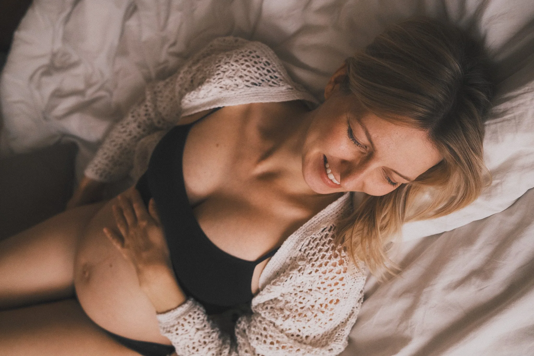 Eine lachende Frau liegt auf einem Bett, trägt schwarze Unterwäsche und eine weiße Strickjacke.