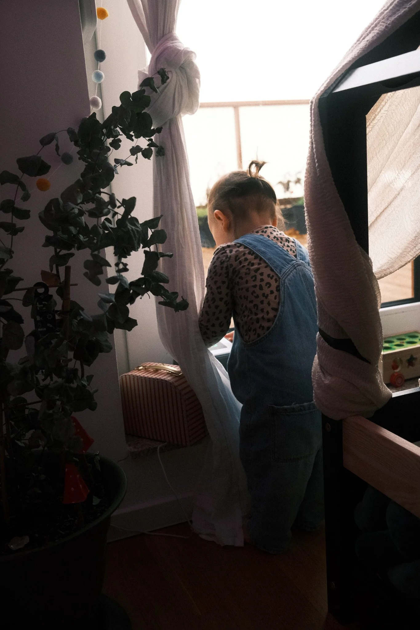 Ein kleines Kind steht am Fenster im warmen Zuhause und schaut nach draußen – ruhige, natürliche Familienfotografie.