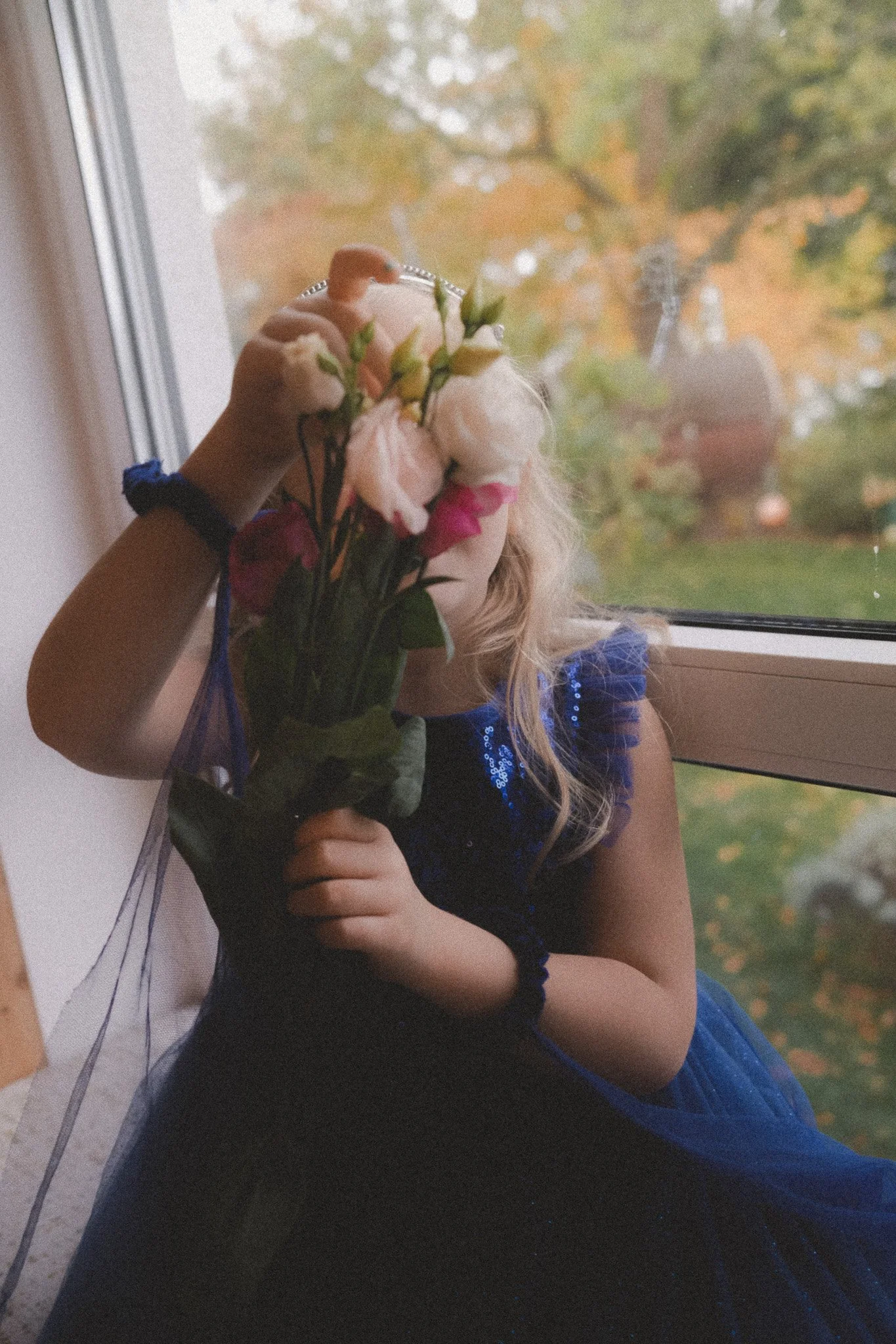 ein Mädchen sitzt am Fenster und hält einen Blumenstrauß vor ihr Gesicht – poetische Kinderfotografie in warmem Herbstlicht.