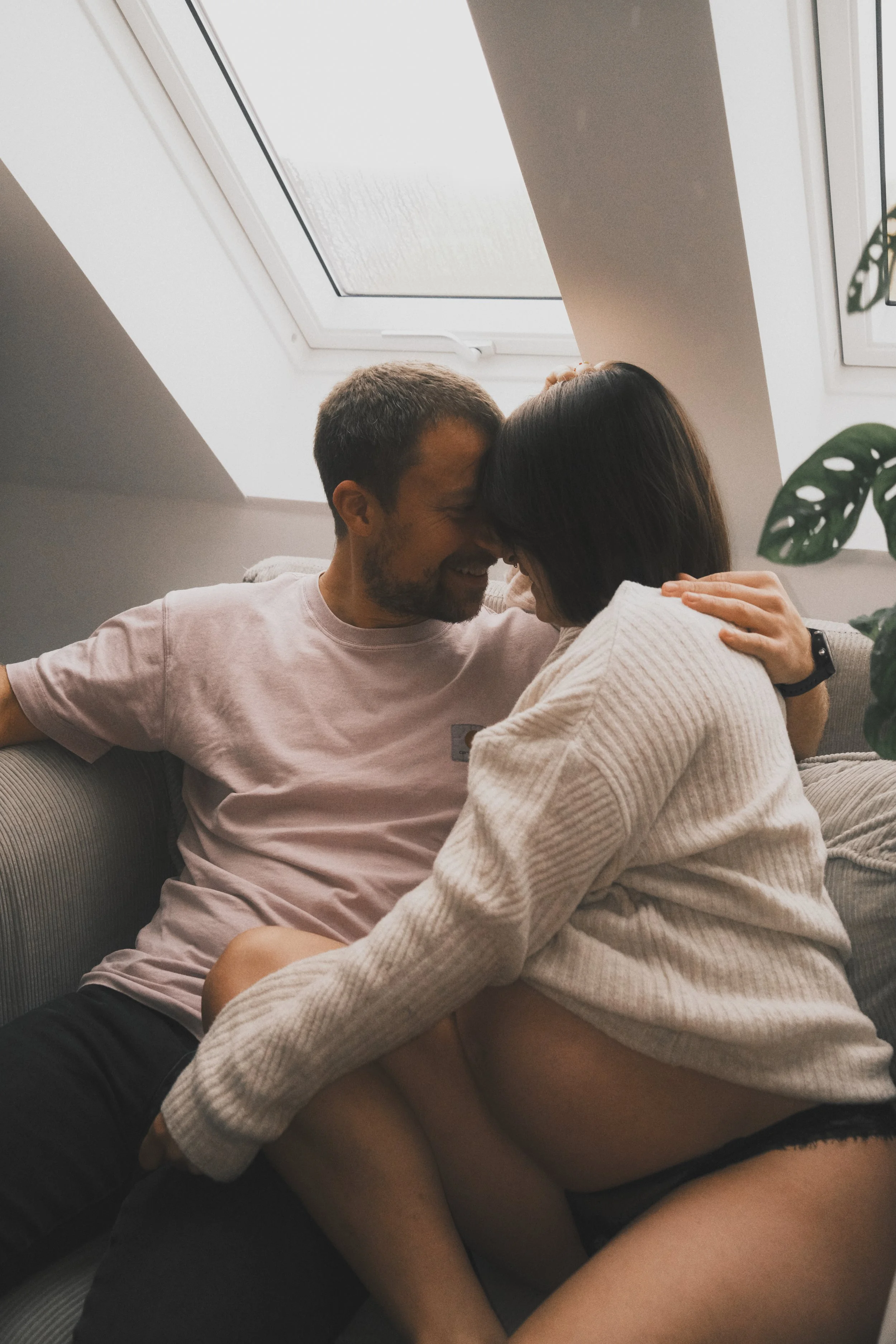Ein glückliches Paar kuschelt auf einem Sofa unter einem Dachfenster, umgeben von Pflanzen.