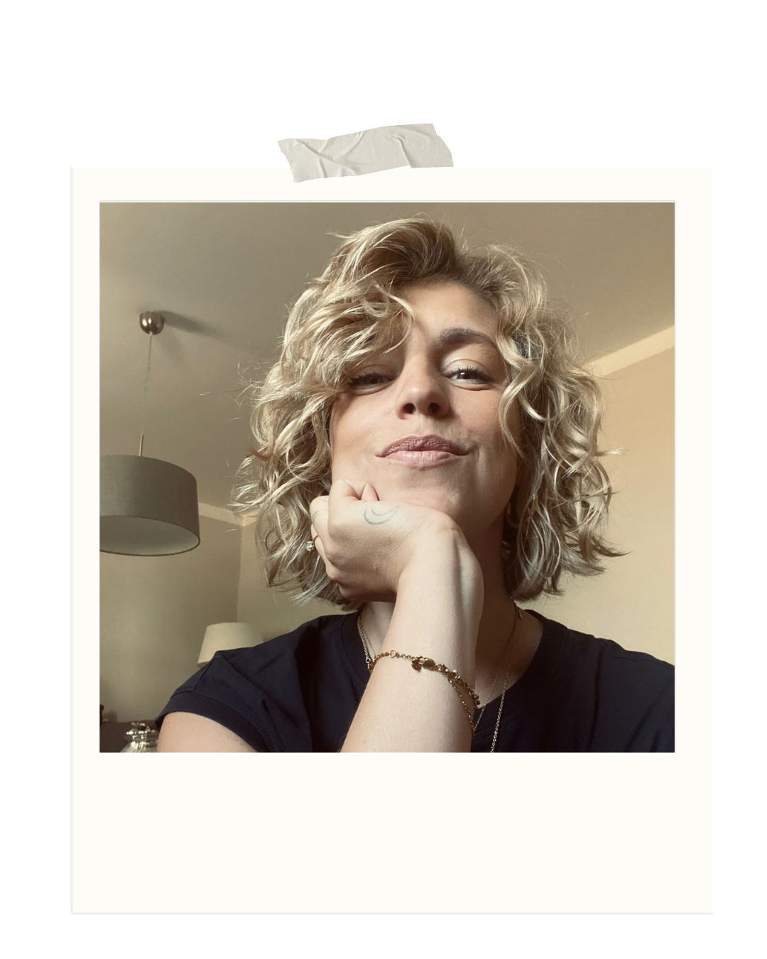 Eine Frau mit lockigem, blonden Haar posiert lächelnd. Sie ruht ihren Kopf auf ihrer Hand, trägt eine schwarze T-Shirt und mehrere Armbänder, sowie einen Ring. Der Hintergrund zeigt einen Raum mit Lampen und hellen Wänden.