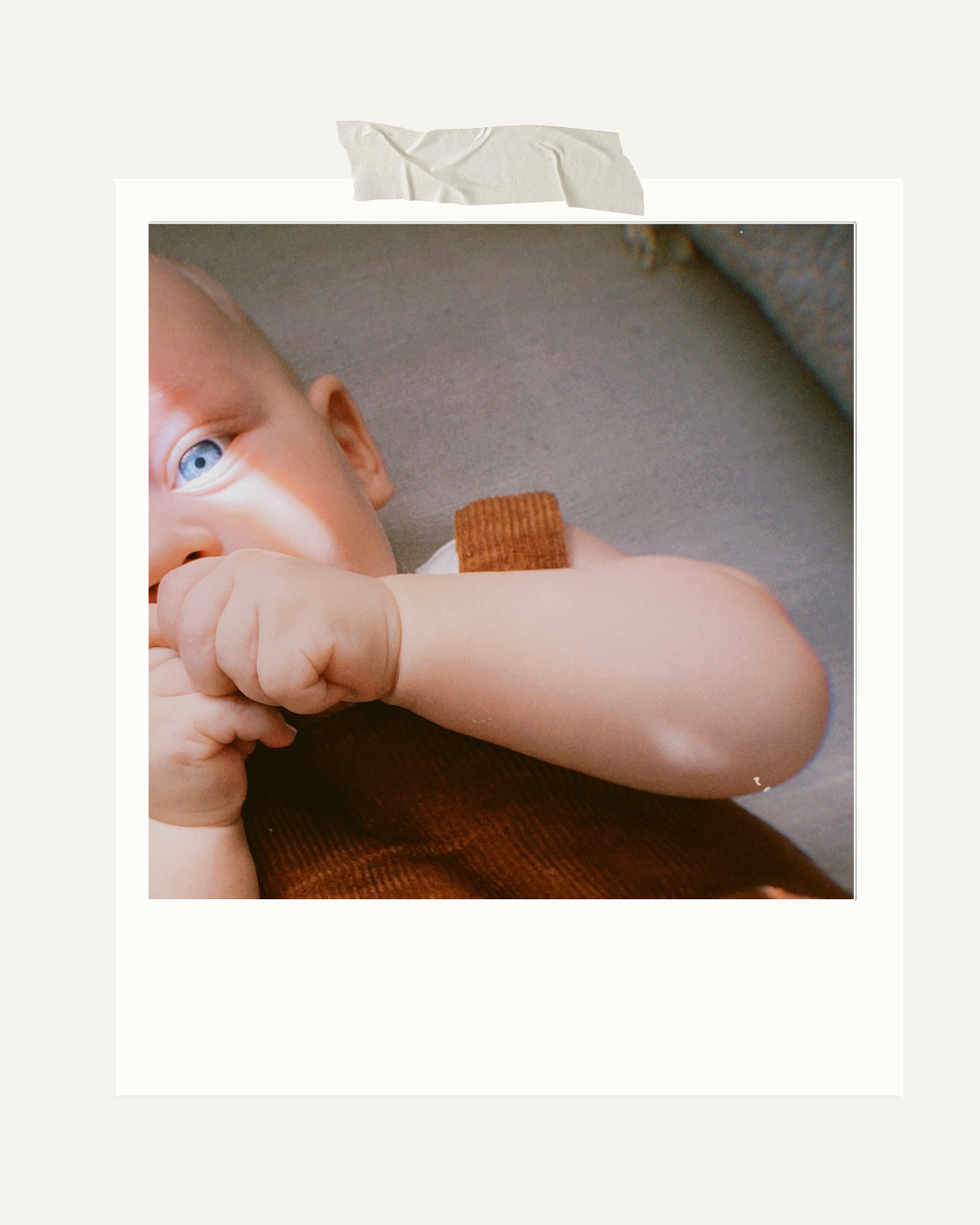 Nahaufnahme eines Babys mit blauen Augen, das auf dem Bauch liegt und seine Hand in den Mund steckt, trägt ein braunes Oberteil.