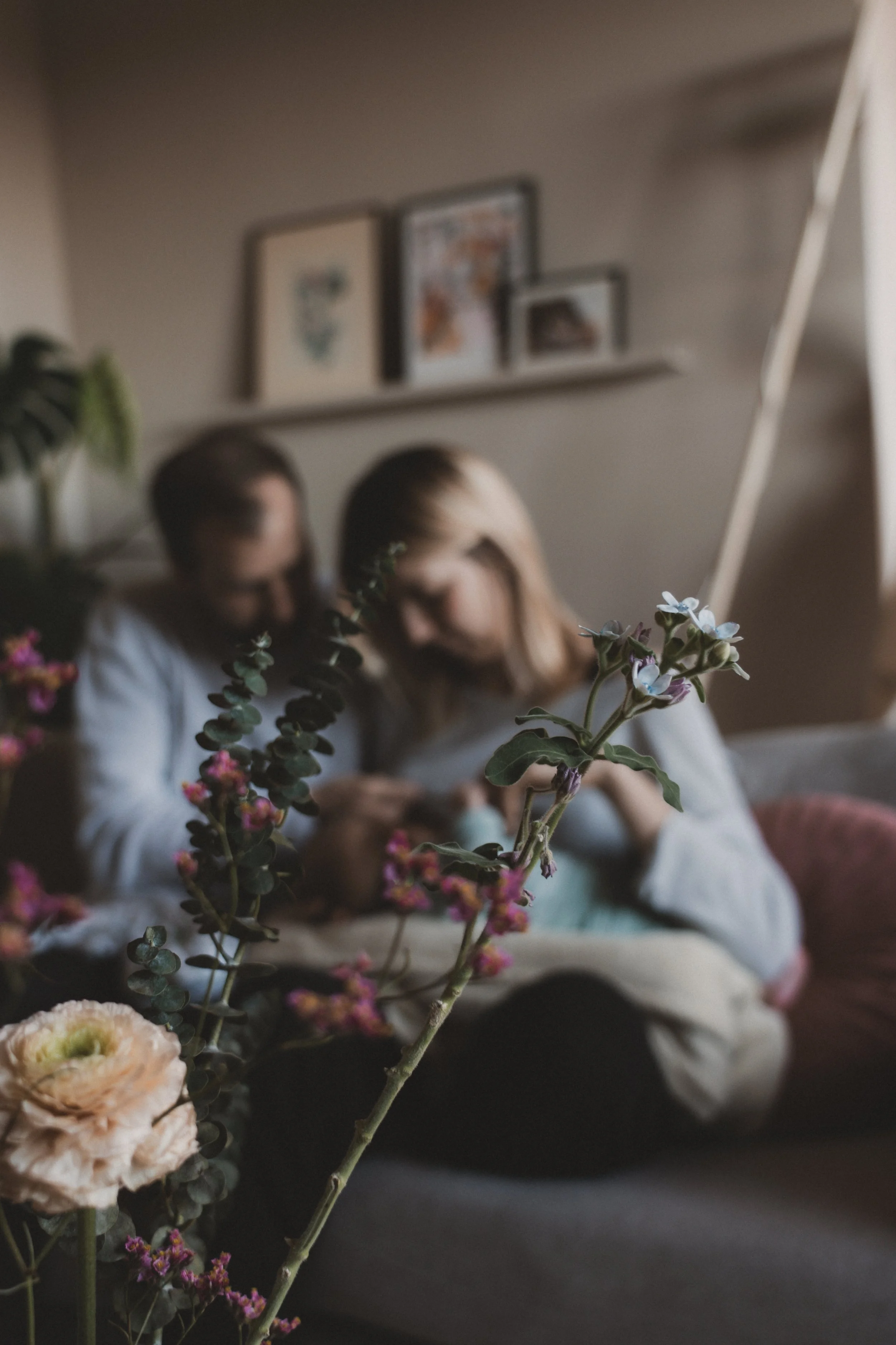 Fokus auf eine Blume im Vordergrund, im Hintergrund zwei Menschen, die auf einem Sofa sitzen und ein Smartphone anschauen, mit unscharfer Inneneinrichtung und Bilder an der Wand.