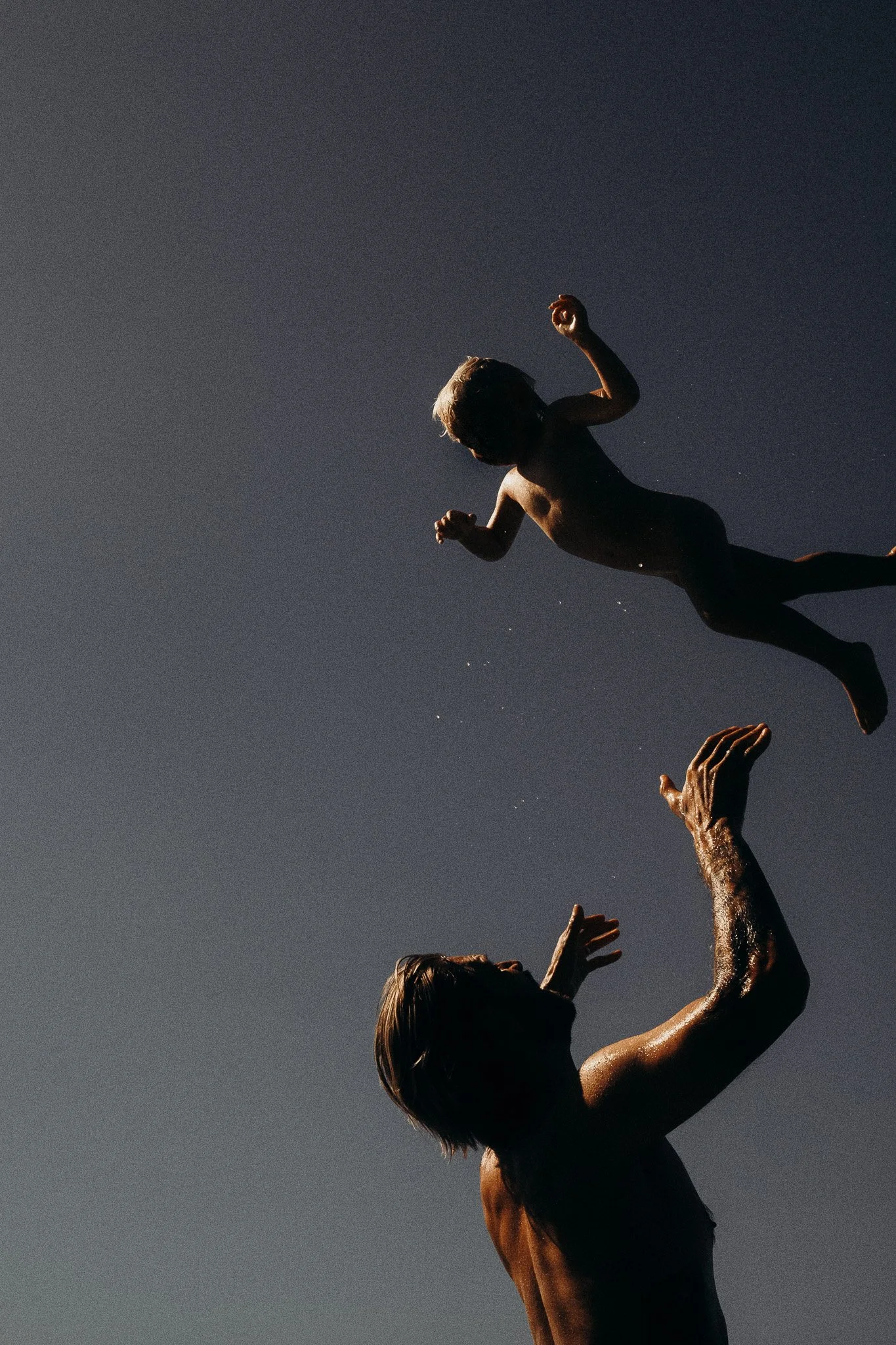 Ein Erwachsener wirft ein Kind in die Luft, beide sind nur in Badebekleidung und stehen im Wasser, der Himmel ist klar.