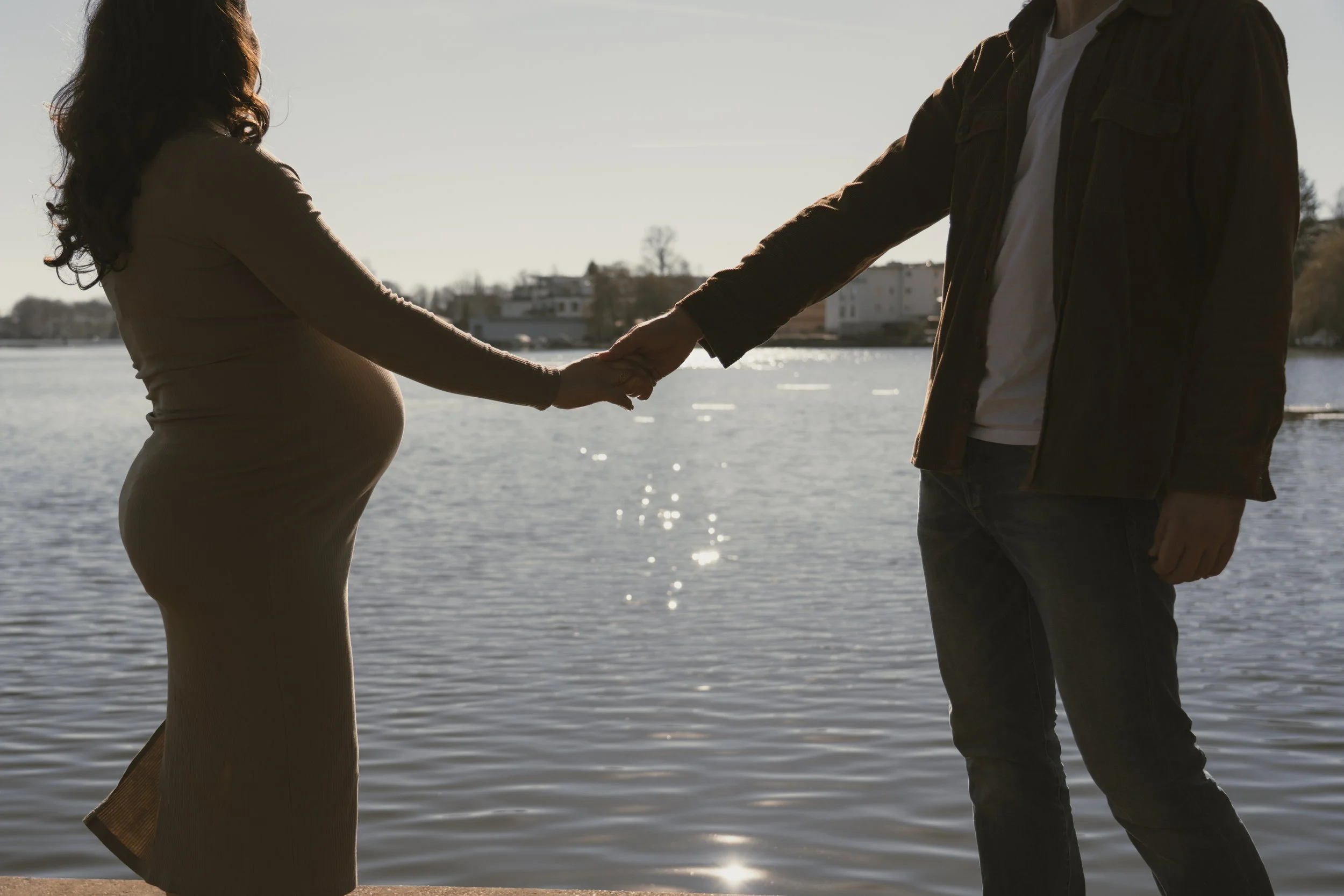 Paar hält Händchen am Flussufer, Frau ist schwanger, Sonnenuntergang im Hintergrund.