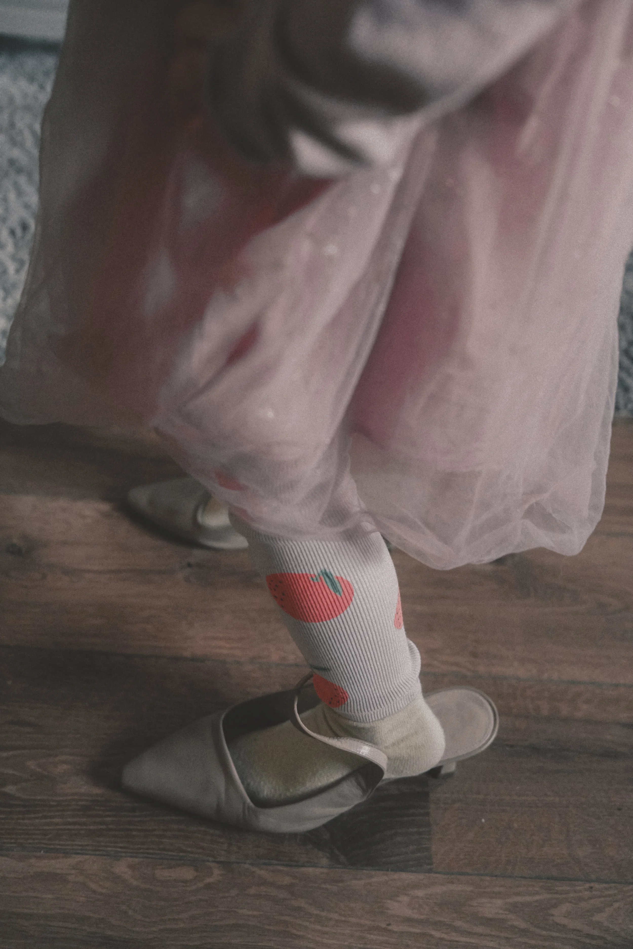 Nahaufnahme eines Beines mit socke und Absatzschuhen, daneben eine rosa, leicht glänzende Tüllrock und ein Teil eines grauen Oberteils, auf dunklem Holzboden