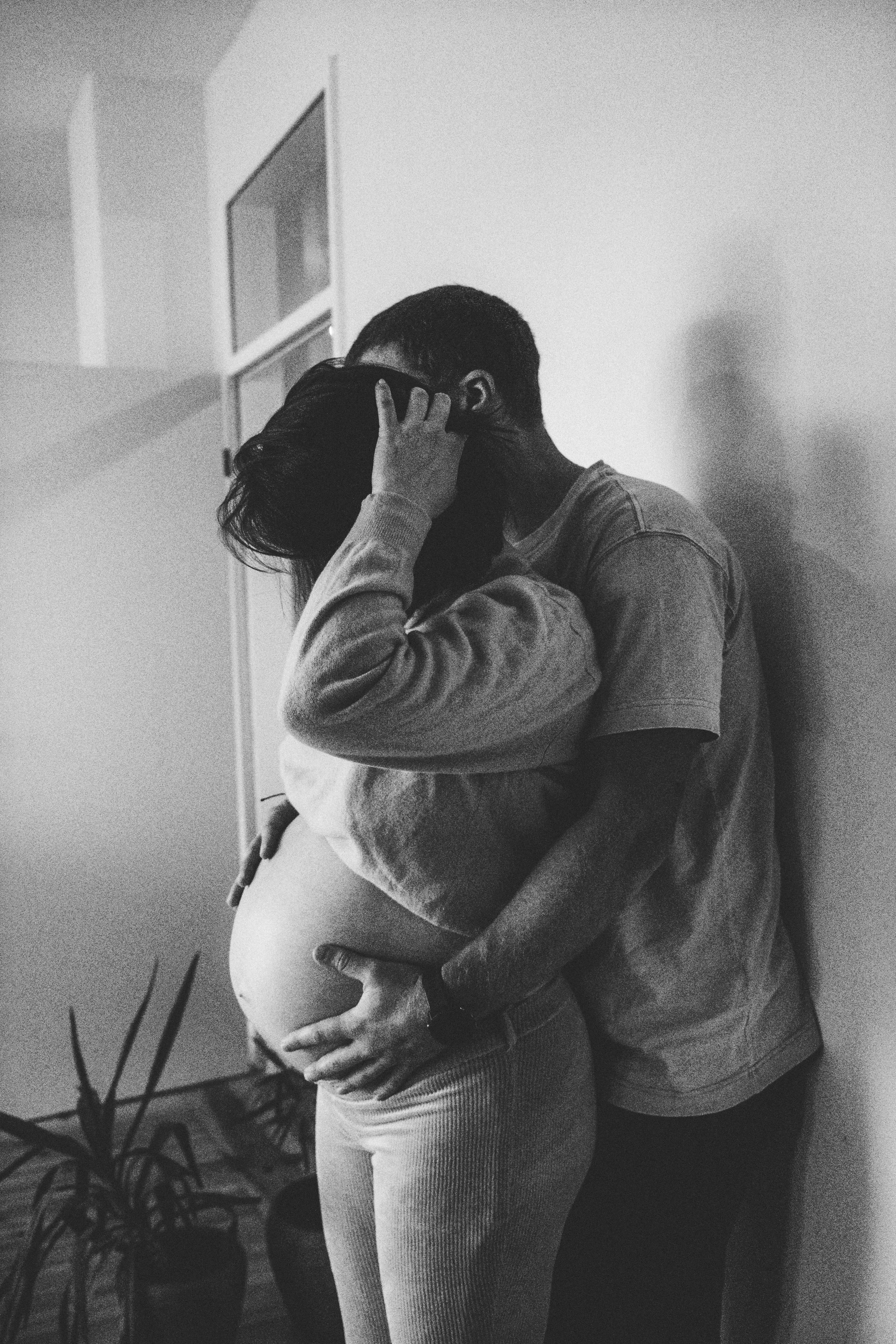 Ein Paar umarmt sich eng, die Frau ist schwanger, das Bild ist schwarz-weiß, im Hintergrund sind Pflanzen und eine Wand.